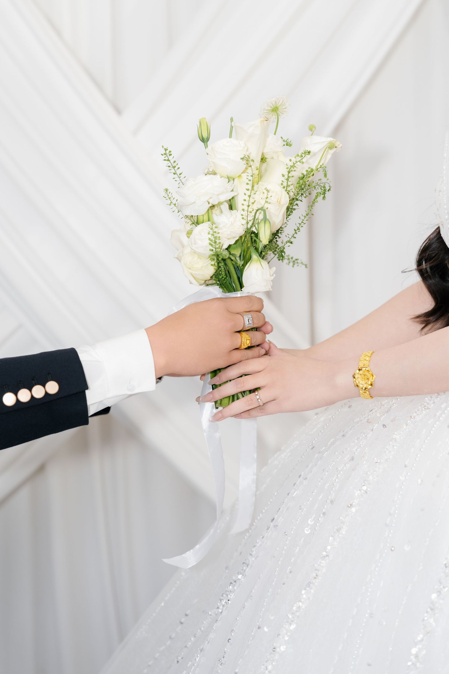 Bride and groom hands exchanging white floral bouquet, bride wearing gold bracelet and diamond rings.