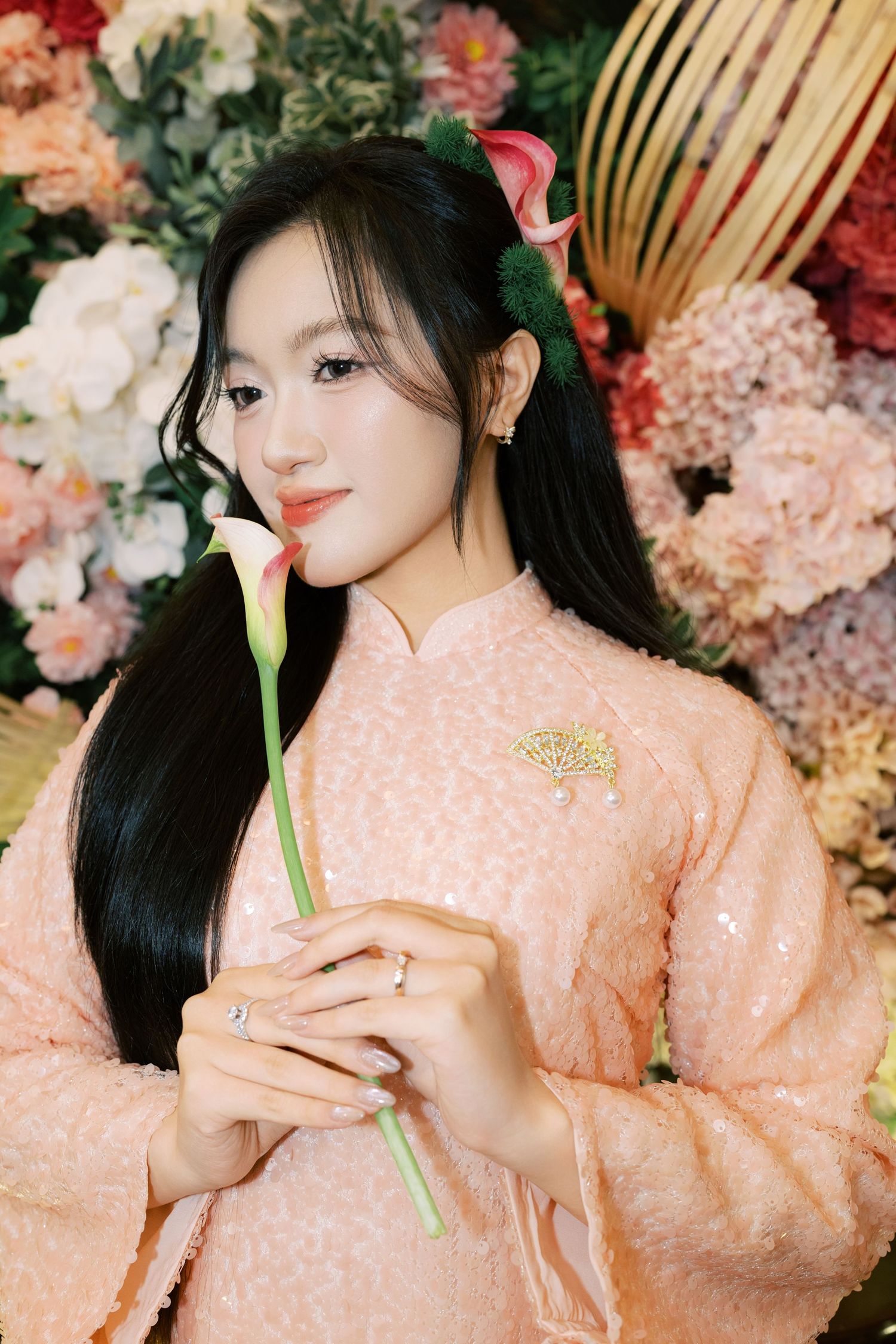 Young Vietnamese woman in pink ao dai holding a calla lily with floral background.