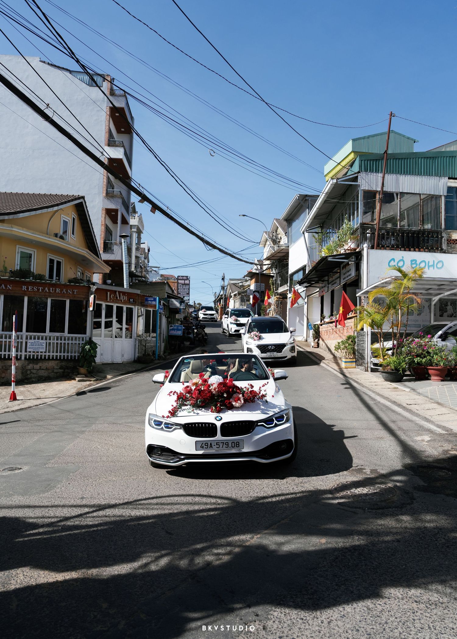 White BMW convertible adorned with red roses drives through a charming Vietnamese street lined with colorful buildings.
