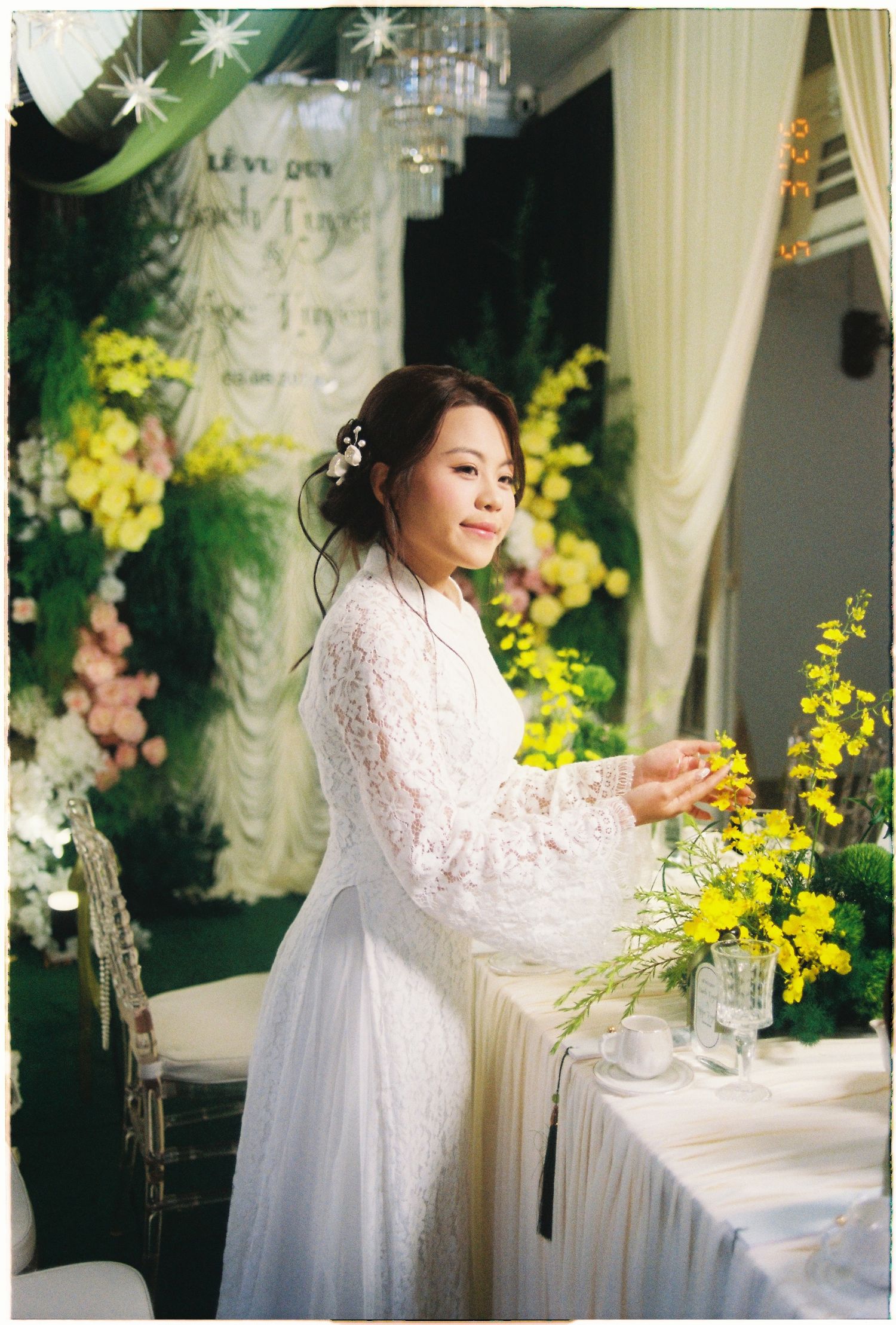 Bride in white lace dress arranging yellow flowers at wedding reception with green floral backdrop.
