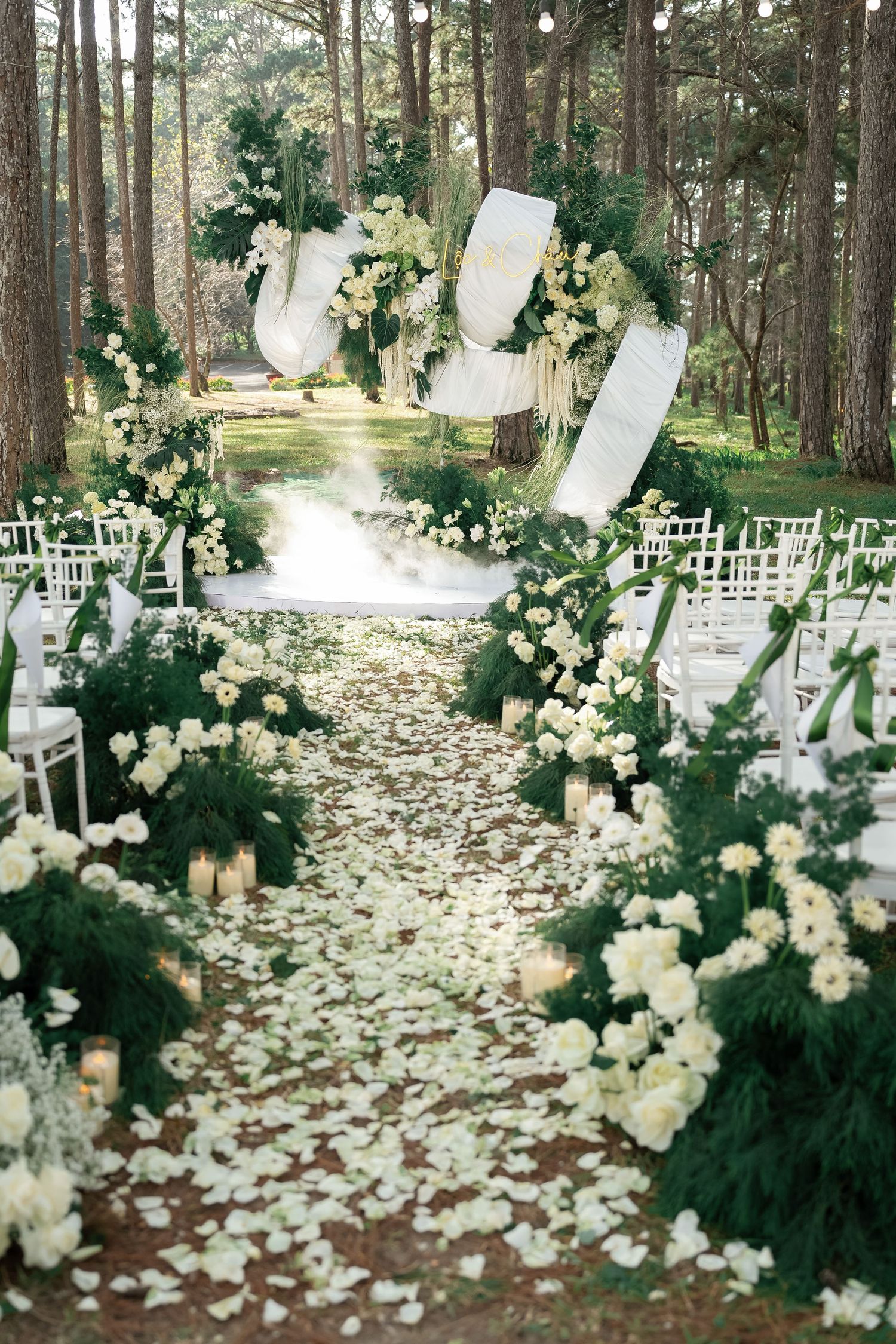 Elegant outdoor wedding aisle lined with white roses, greenery, candles, and flowing white drapes among tall pine trees.