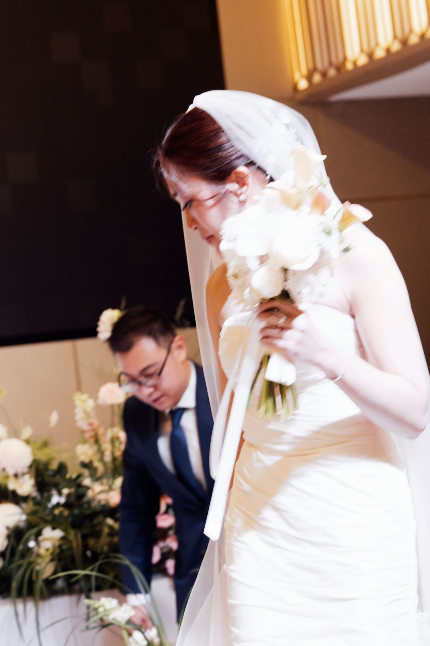Bride in white wedding dress and veil holds bouquet of white flowers while groom signs documents at ceremony.