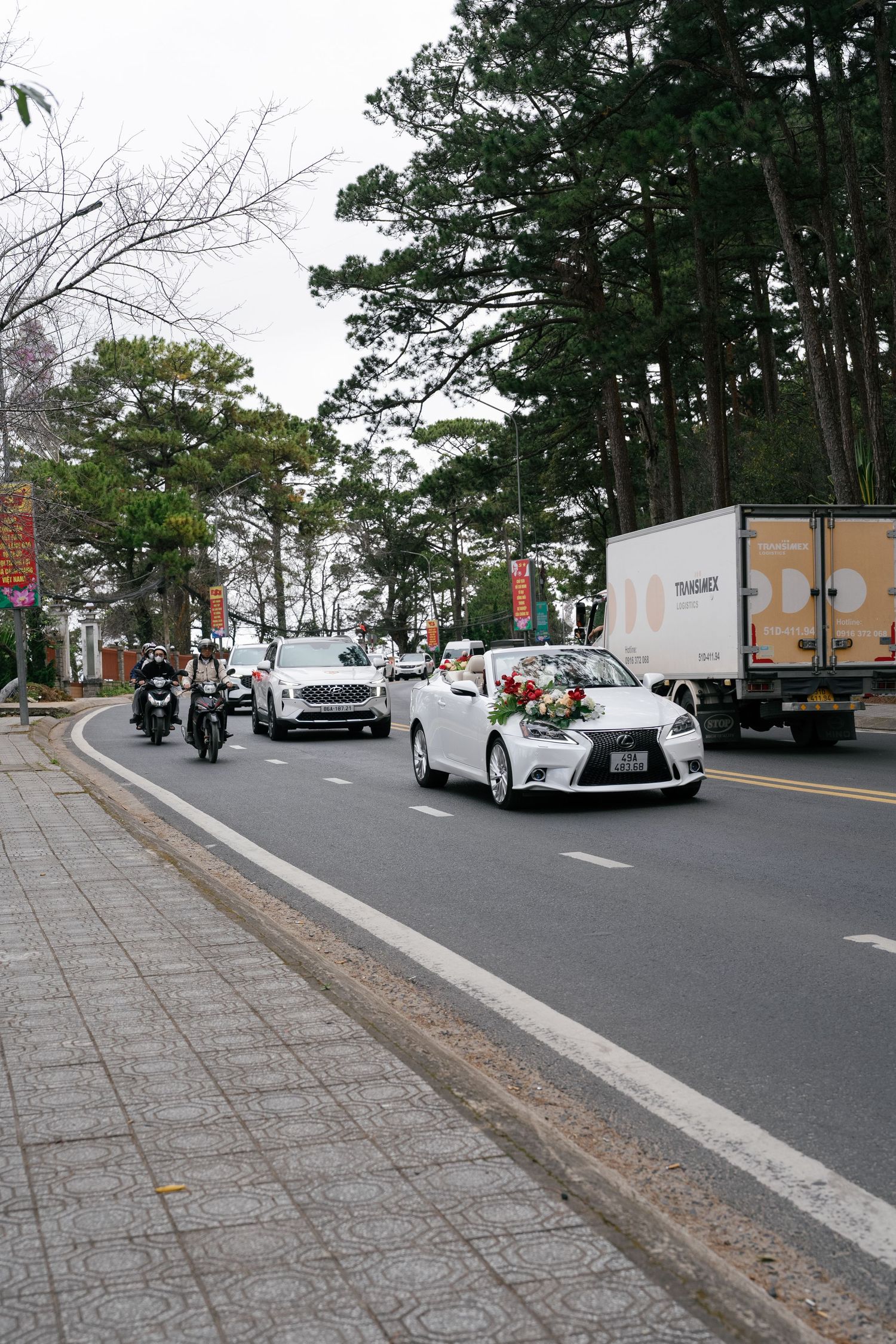 White Lexus wedding car decorated with flowers leads a bridal convoy along a tree-lined road in Vietnam.