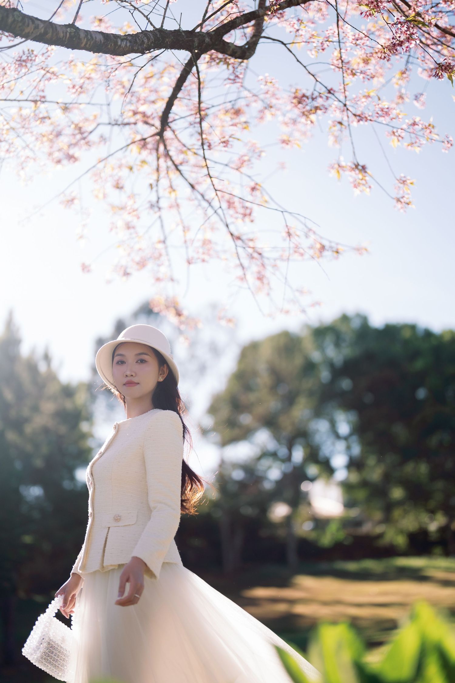 Young woman in white outfit and hat standing under blooming cherry blossom tree in sunlit park.