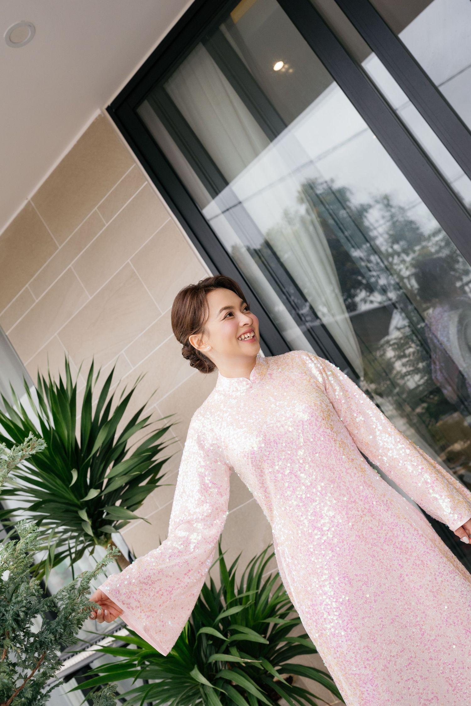 A smiling woman in a pink sequined long-sleeve dress poses near tropical plants and large windows.