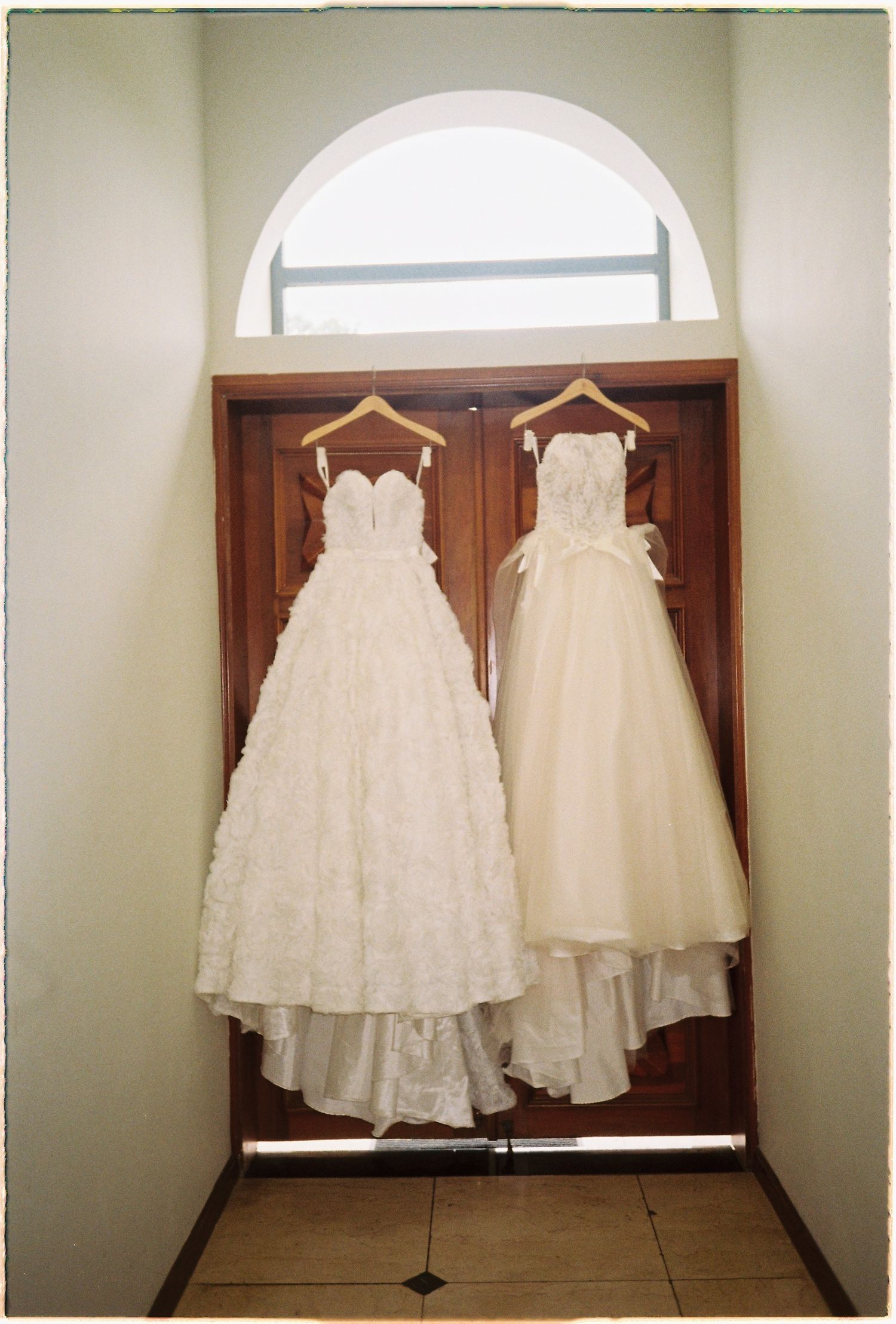 Two elegant white wedding gowns hanging on wooden hangers against a rustic wooden door with arched window above.