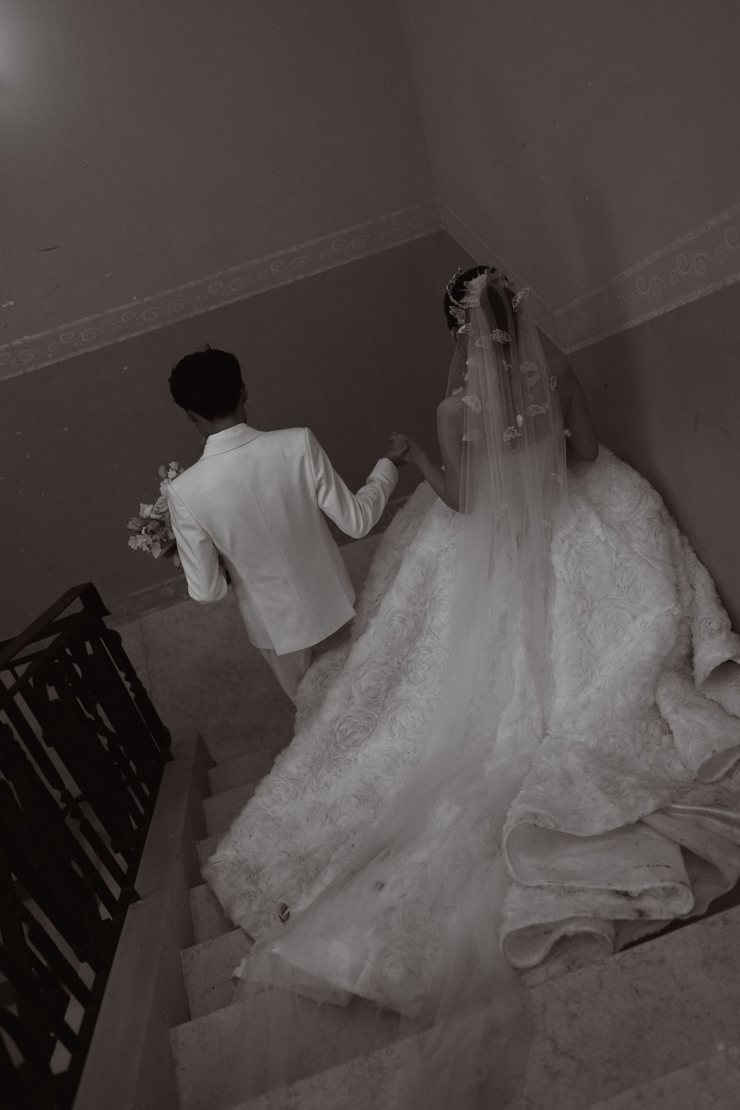 Black and white photo of bride and groom holding hands walking up stairs, bride wearing lace gown and veil.
