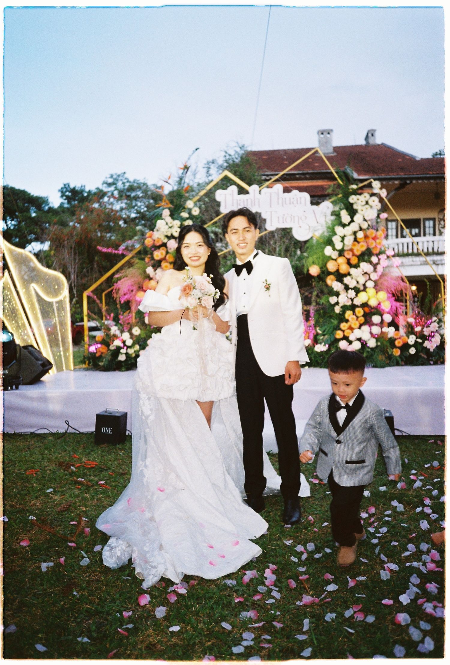 Bride and groom in white wedding attire pose with young boy in suit at outdoor floral ceremony.