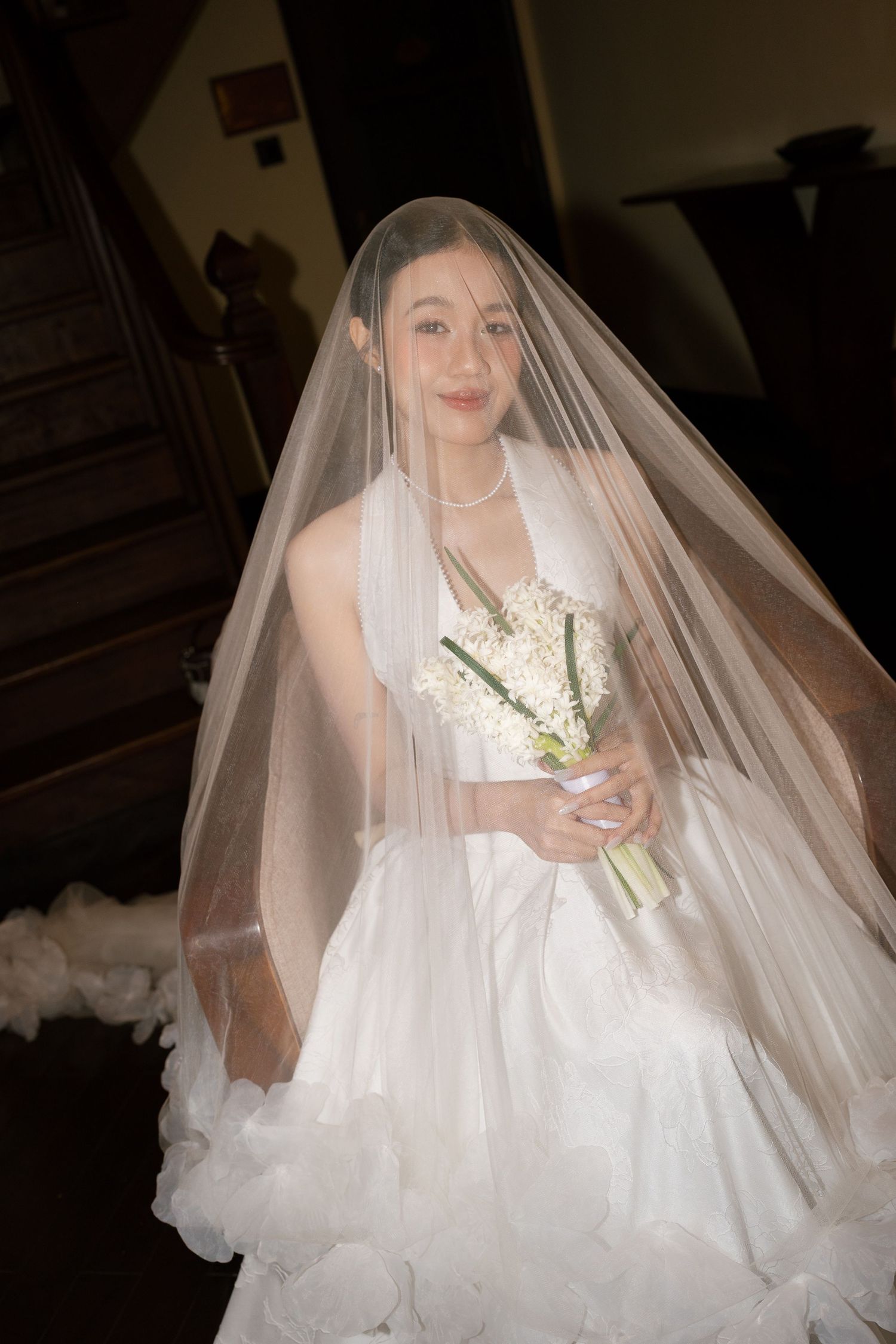 Smiling bride in white gown and veil holding white bouquet while seated on wooden staircase.