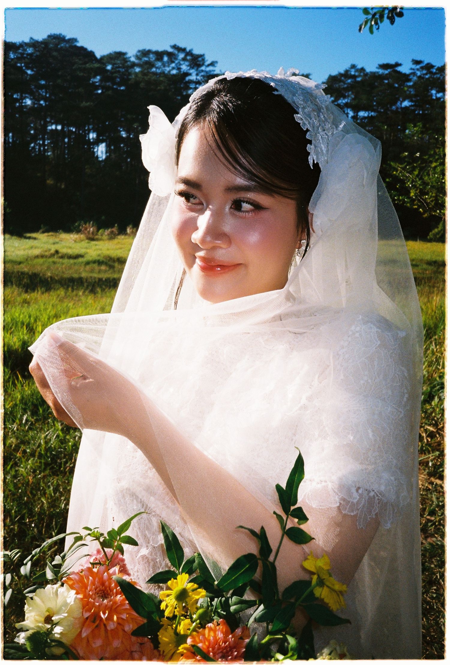 Smiling bride in white lace dress and veil holds colorful flowers in a sunny green meadow setting.
