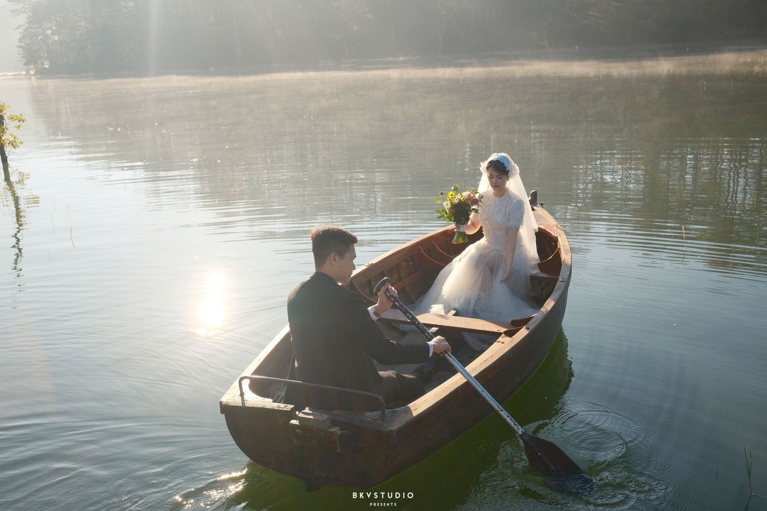 Bride and groom rowing a wooden boat on a sunlit lake, with golden morning light reflecting on the water.