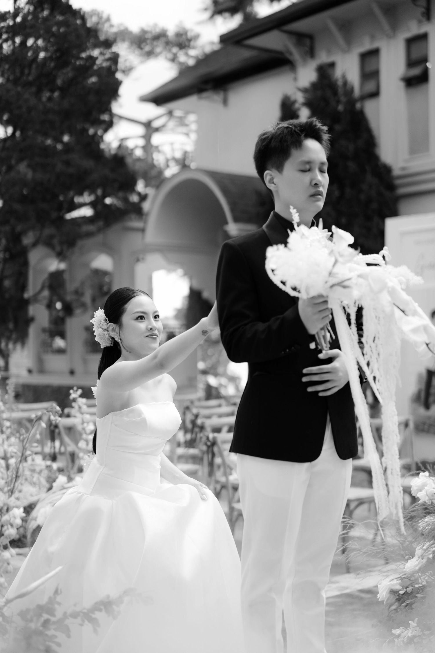 Black and white photo of bride and groom posing outdoors at wedding ceremony with floral bouquet.