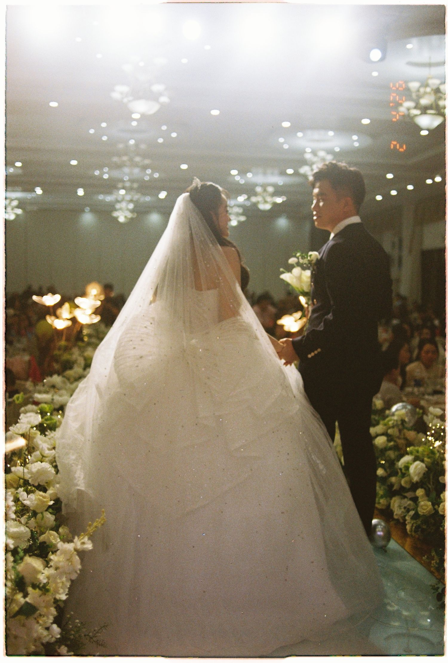 Bride and groom hold hands at altar surrounded by floral arrangements and wedding guests.
