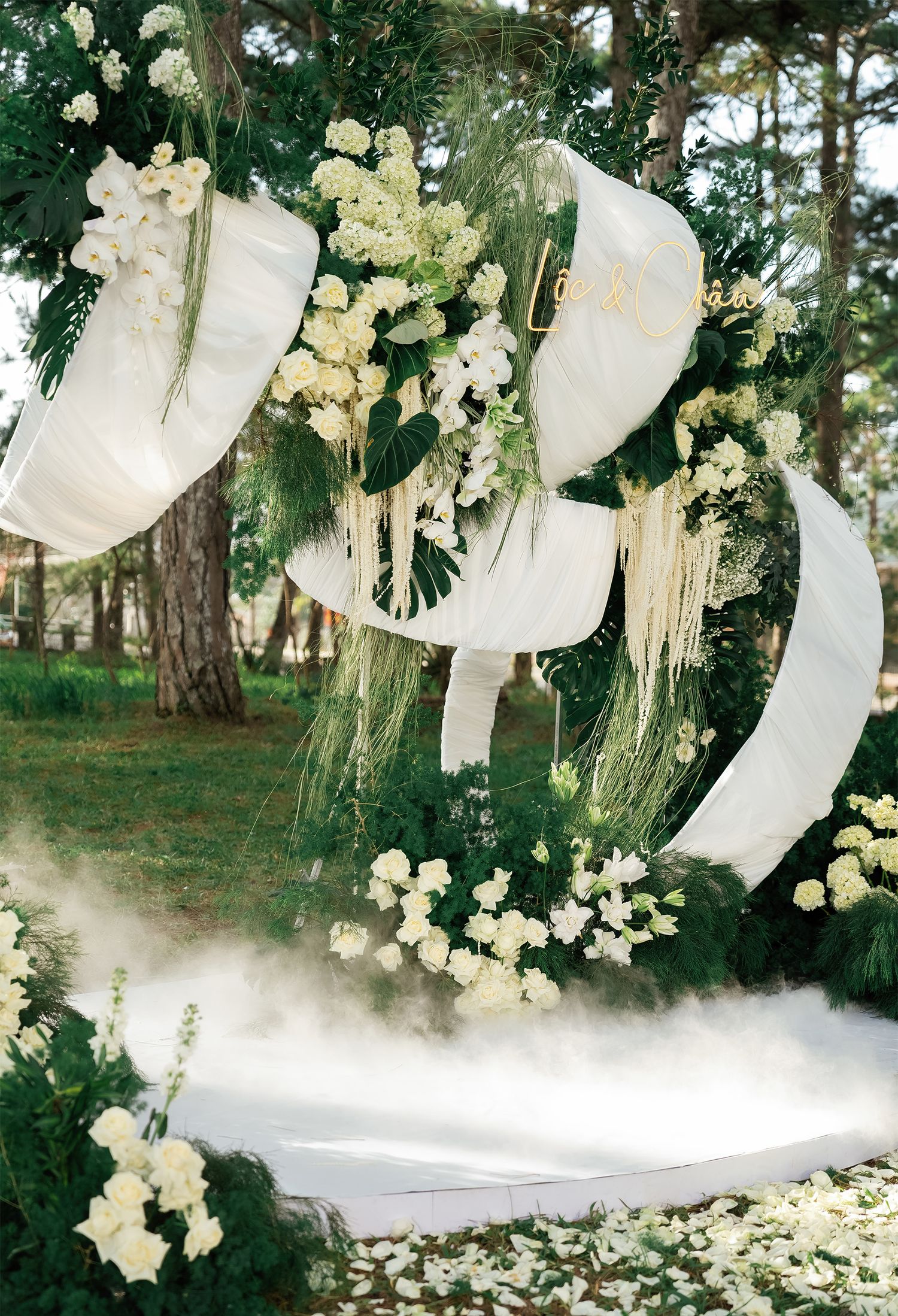 Elegant outdoor wedding arch adorned with white draping, lush greenery, and white floral arrangements amid tall pine trees.