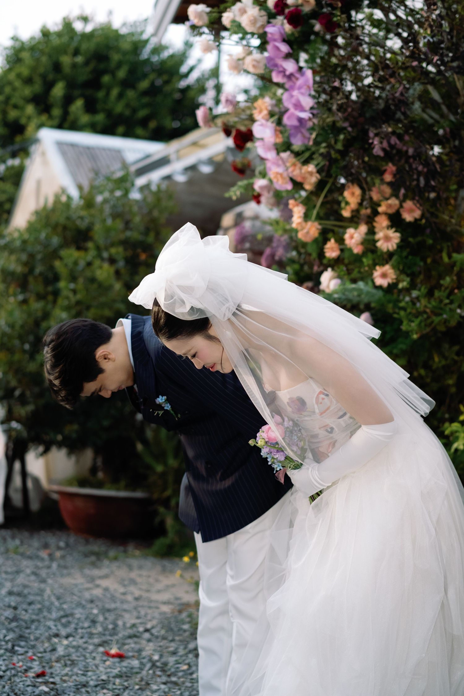 A bride in a white gown and veil shares a tender moment with her groom outdoors near blooming floral arrangements.