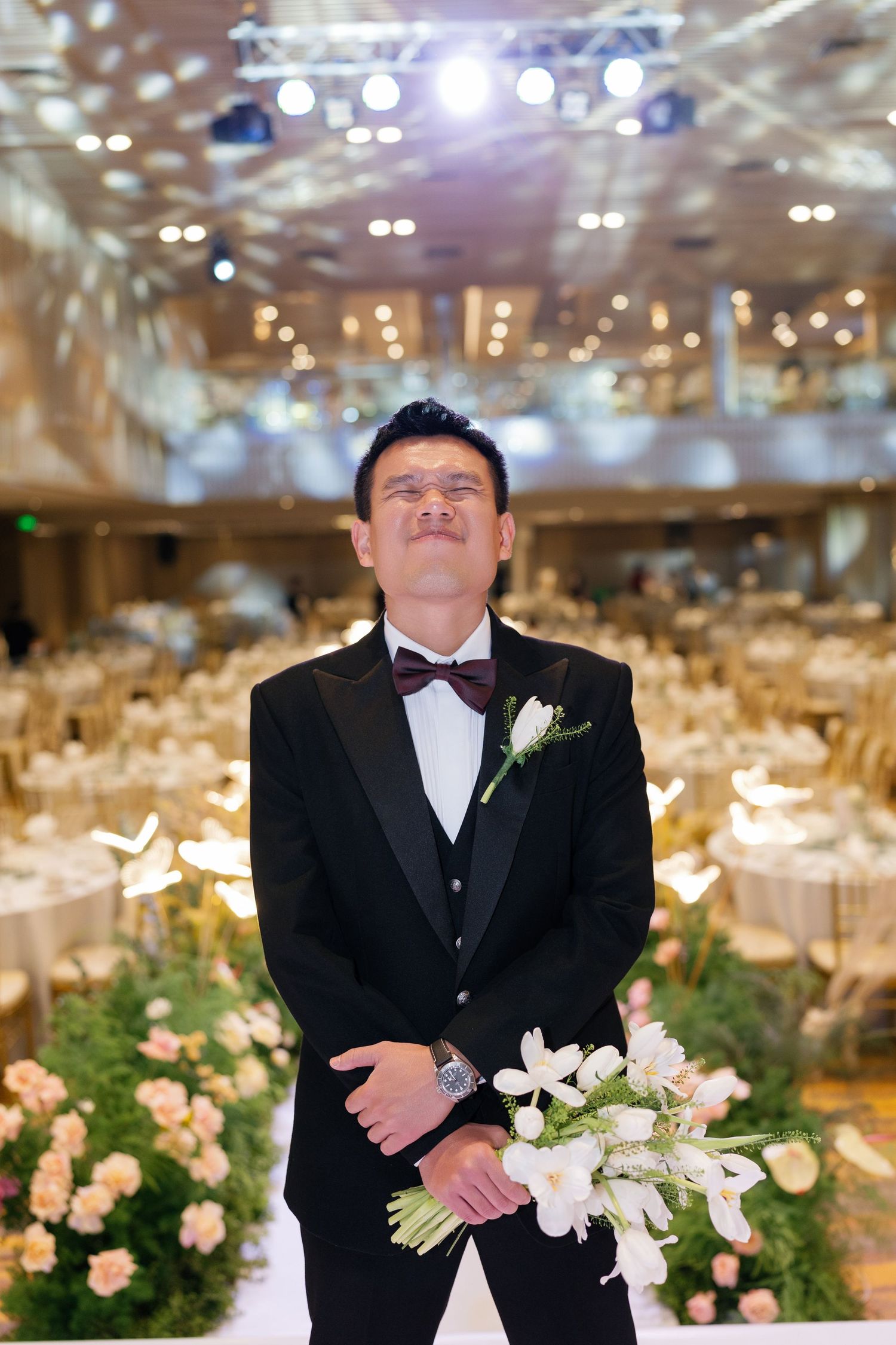 Groom in black tuxedo holding white bouquet standing in elegantly decorated wedding reception hall.