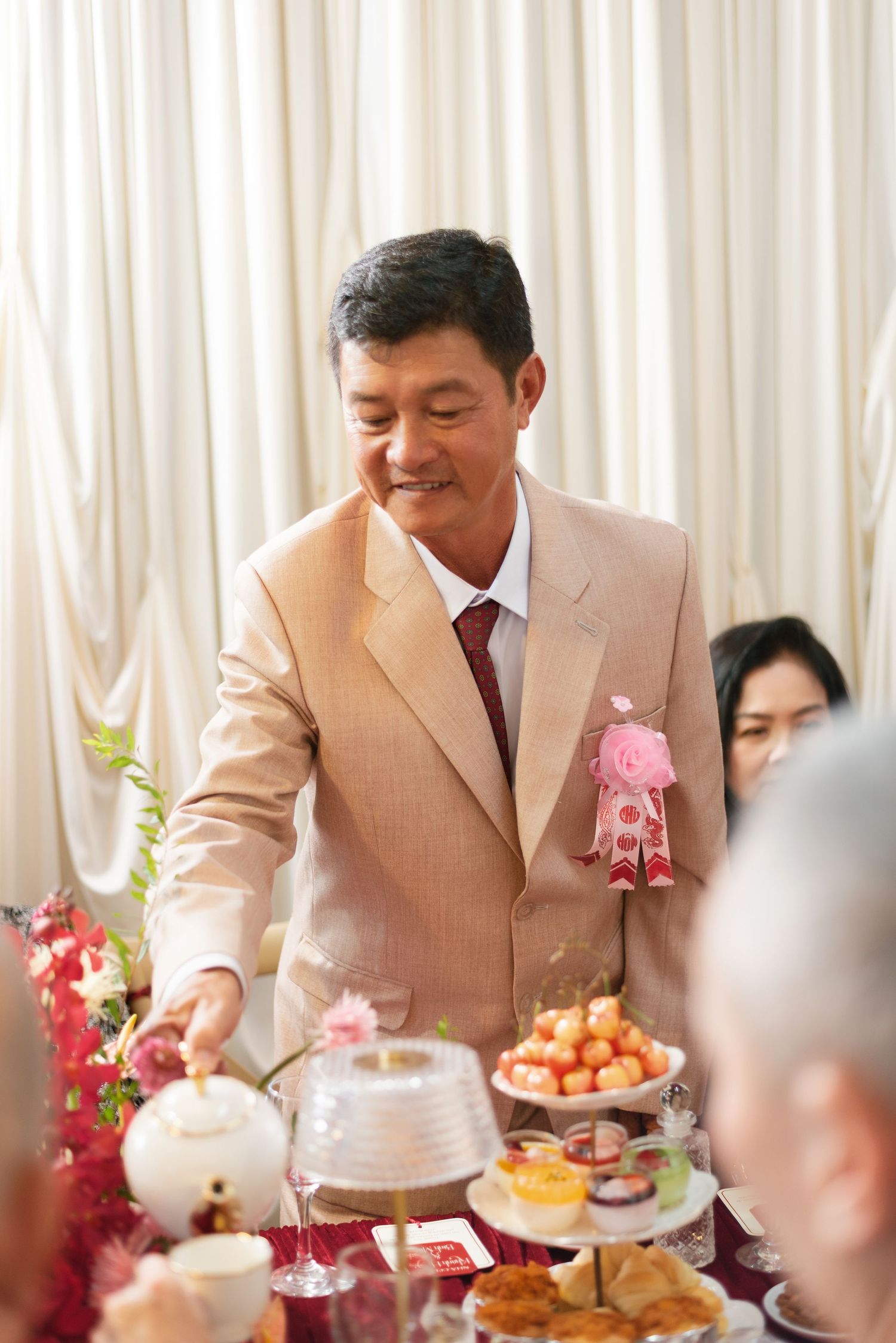 Smiling man in beige suit with pink floral pin serves tea at a traditional Chinese wedding banquet table.
