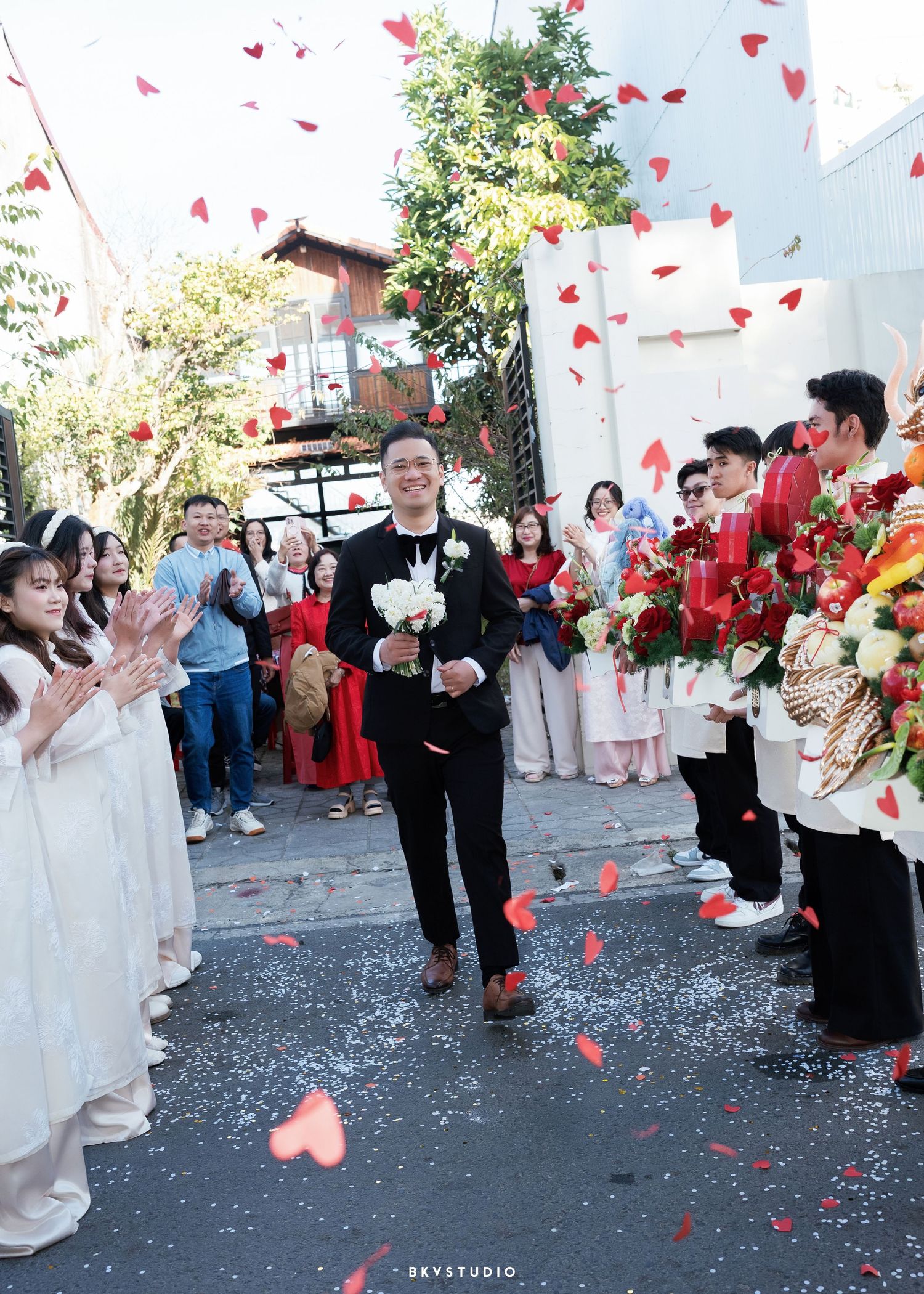 Groom walks through heart confetti shower at Vietnamese wedding ceremony with guests applauding in traditional ao dai attire.