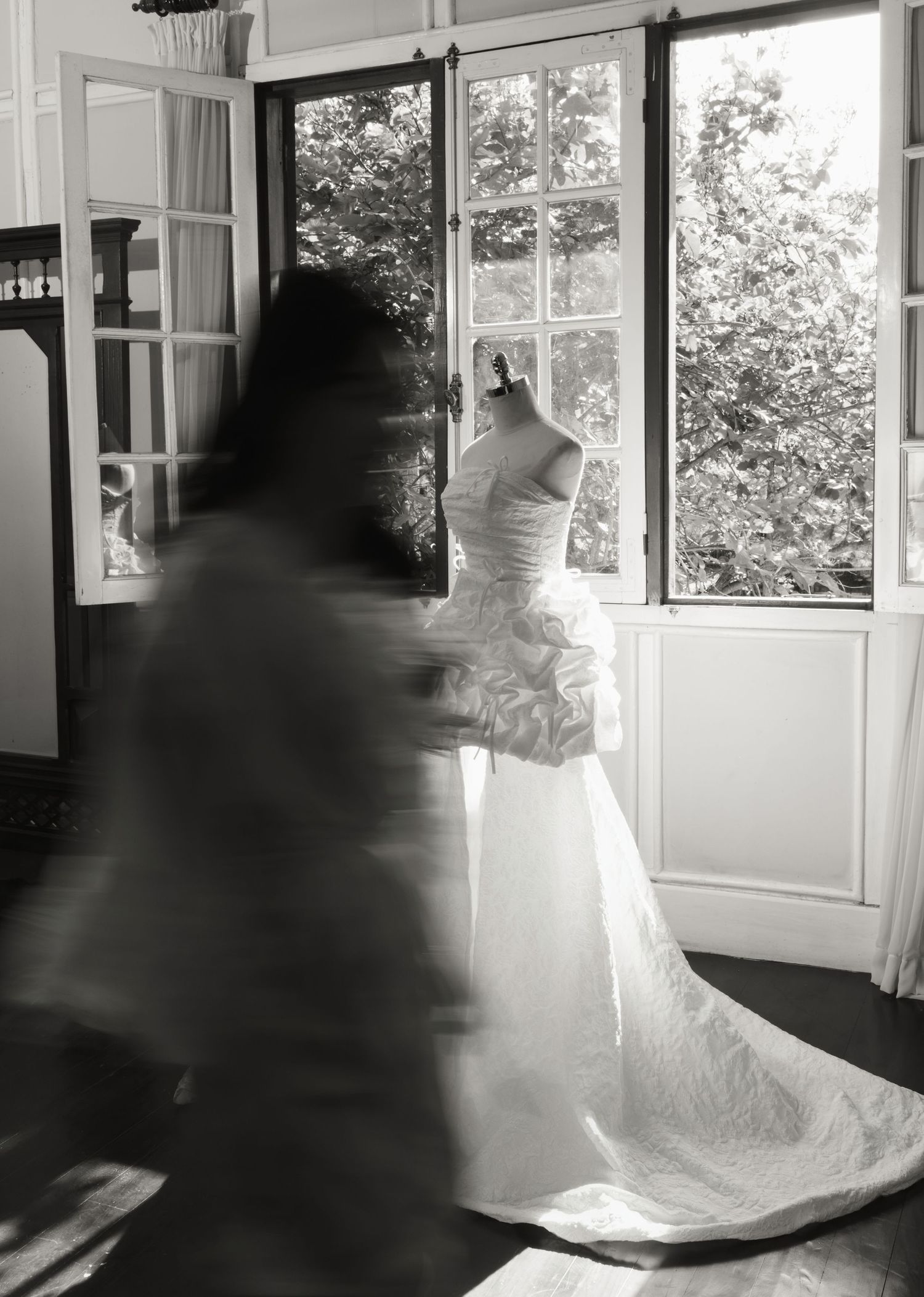 Black and white photo of a wedding dress on a mannequin near French doors with a blurred figure in motion nearby.