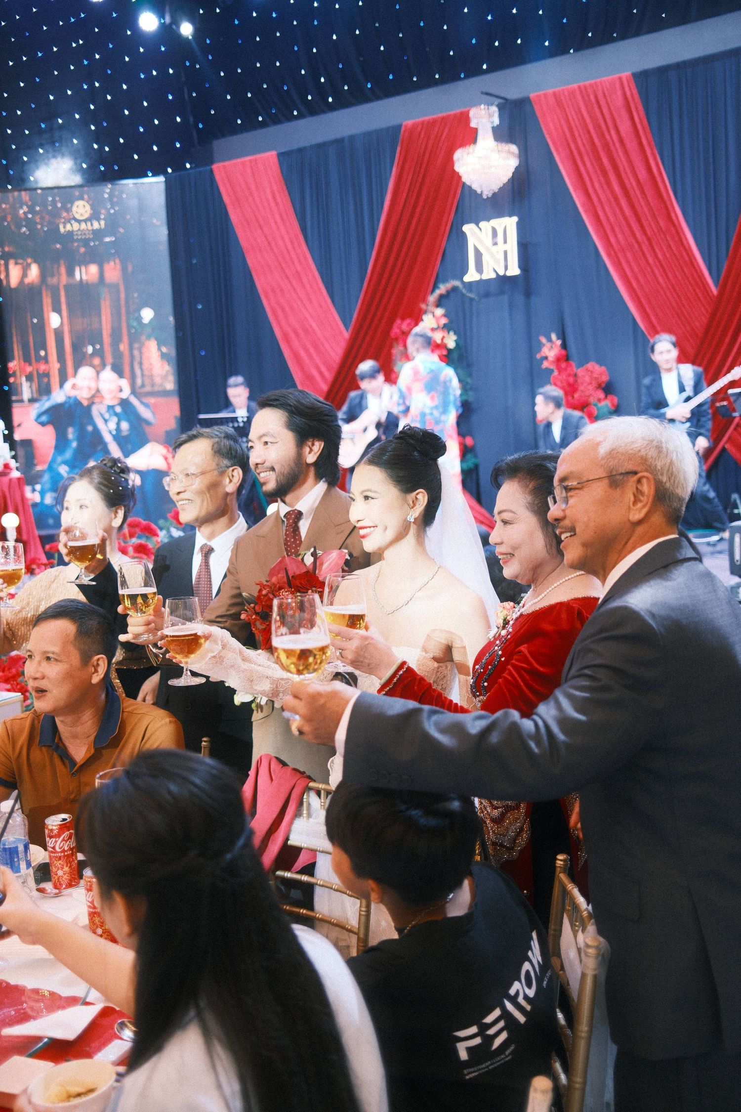Wedding reception toast with bride, groom, and family raising champagne glasses before red draped stage backdrop.