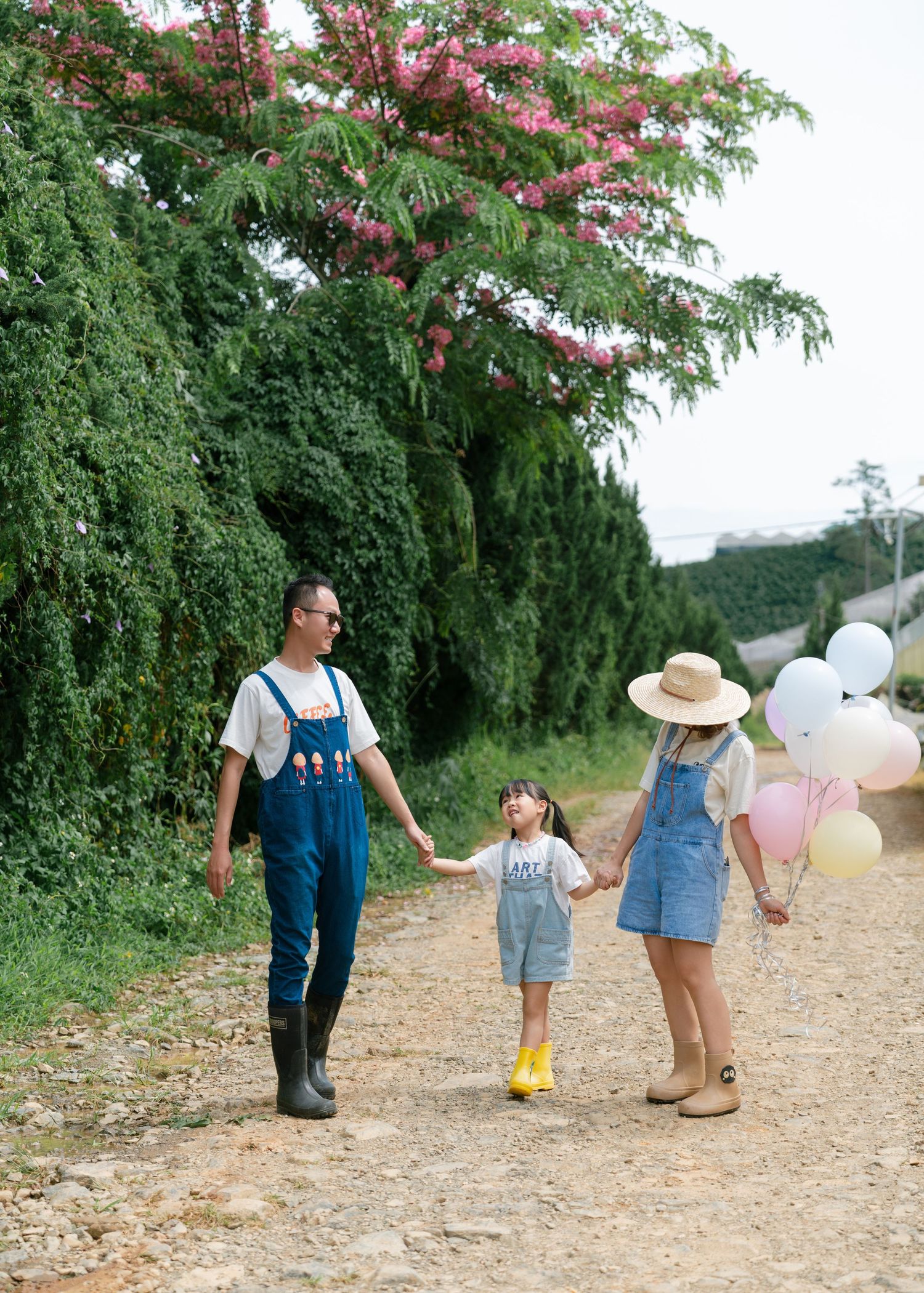 Family of three walking on a dirt path holding hands, with colorful balloons and blooming pink trees in the background.