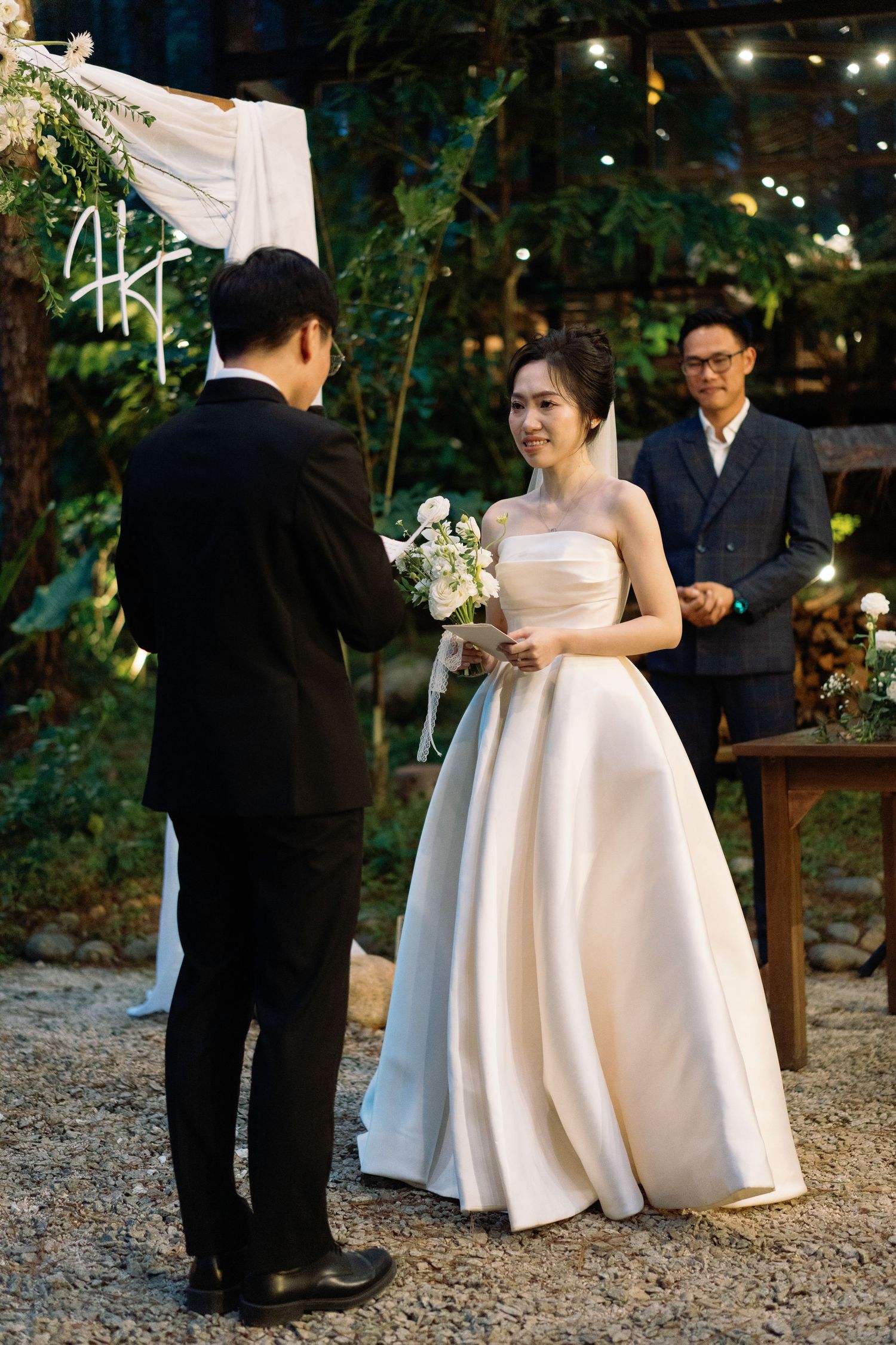 Smiling Asian bride in strapless white ball gown holds bouquet during outdoor wedding ceremony.