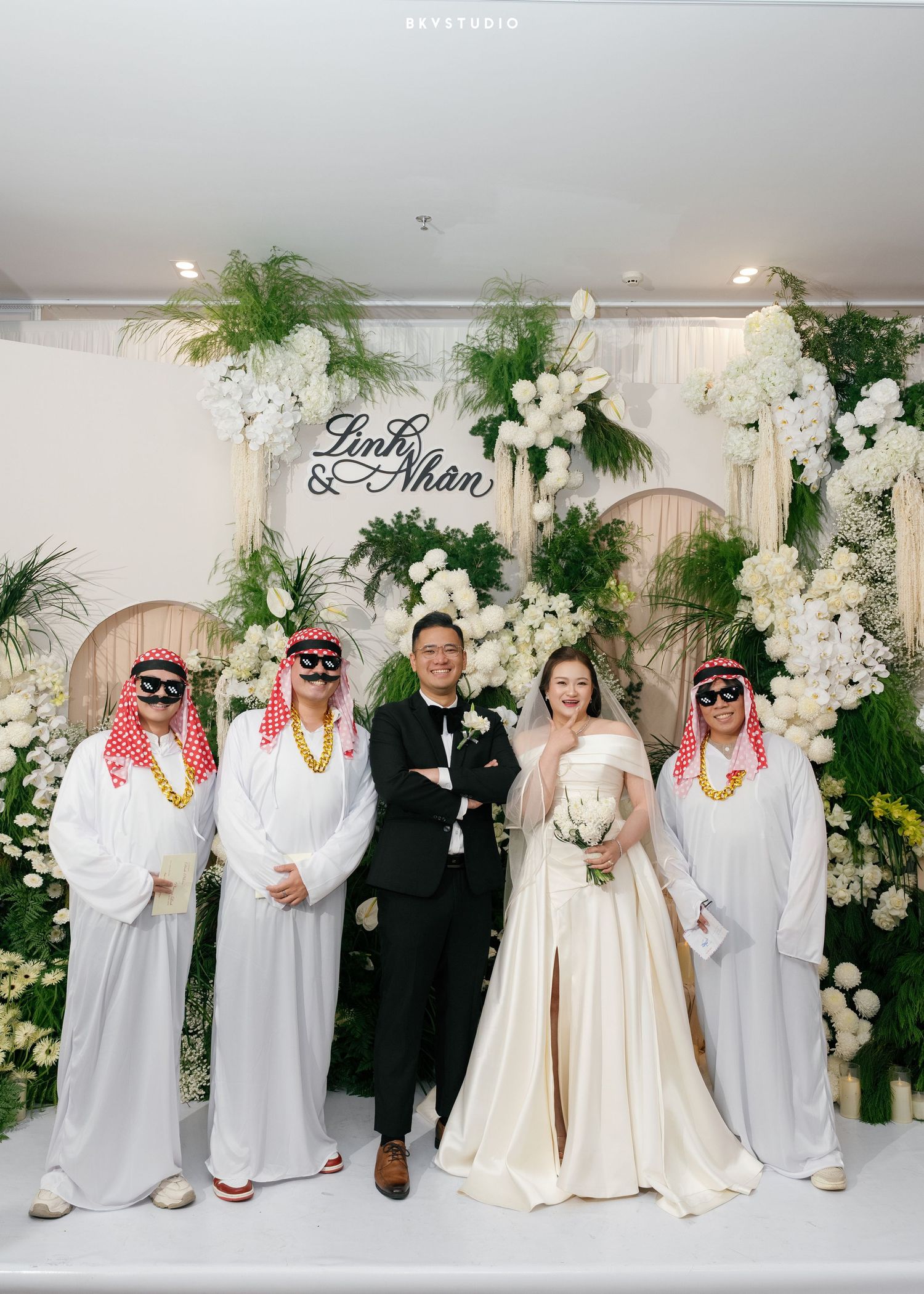 Bride and groom pose with guests in Arab costumes at elegant white floral wedding backdrop reading Linh & Nhan.