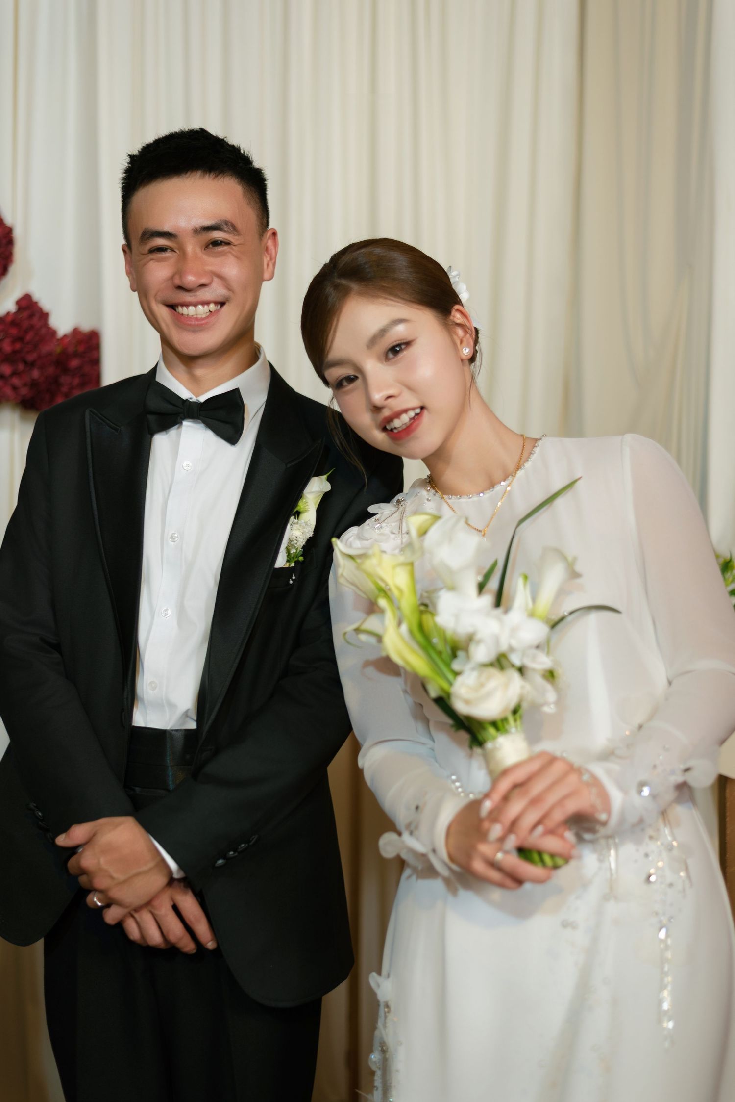 A smiling groom in a black tuxedo stands beside a bride in a white dress holding a bouquet of white calla lilies.