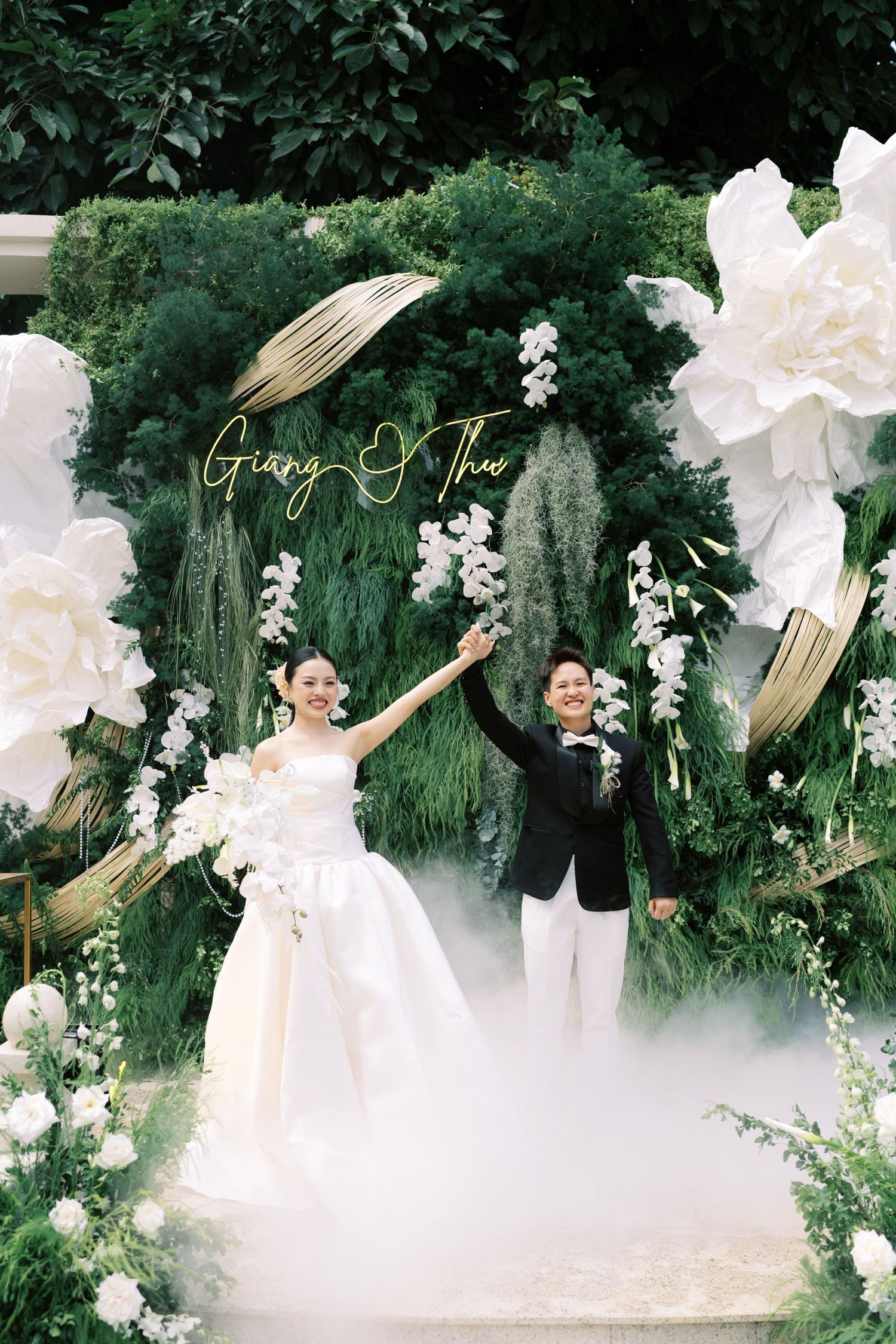 Bride and groom celebrate at green floral arch with giant white flowers and neon name sign.