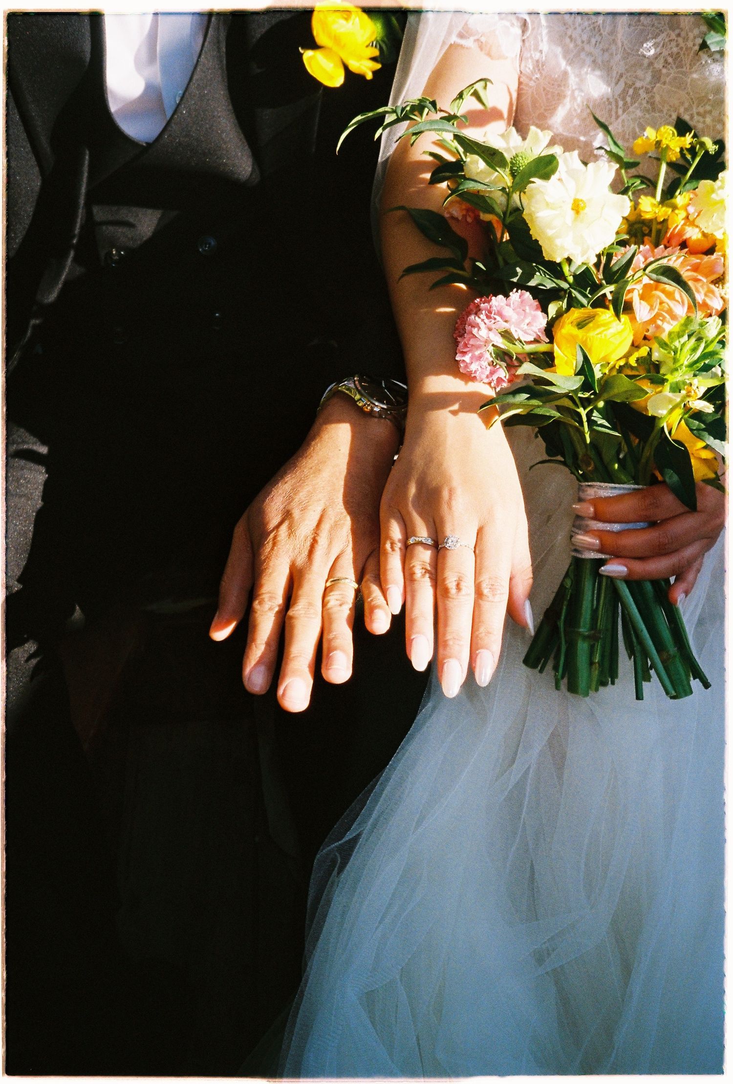 Close-up of bride and groom showing wedding rings while bride holds a vibrant yellow and pink bouquet.