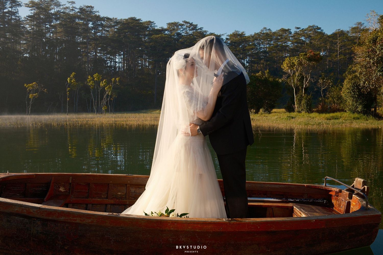 Bride and groom share a kiss on a wooden boat on a serene lake surrounded by lush trees.