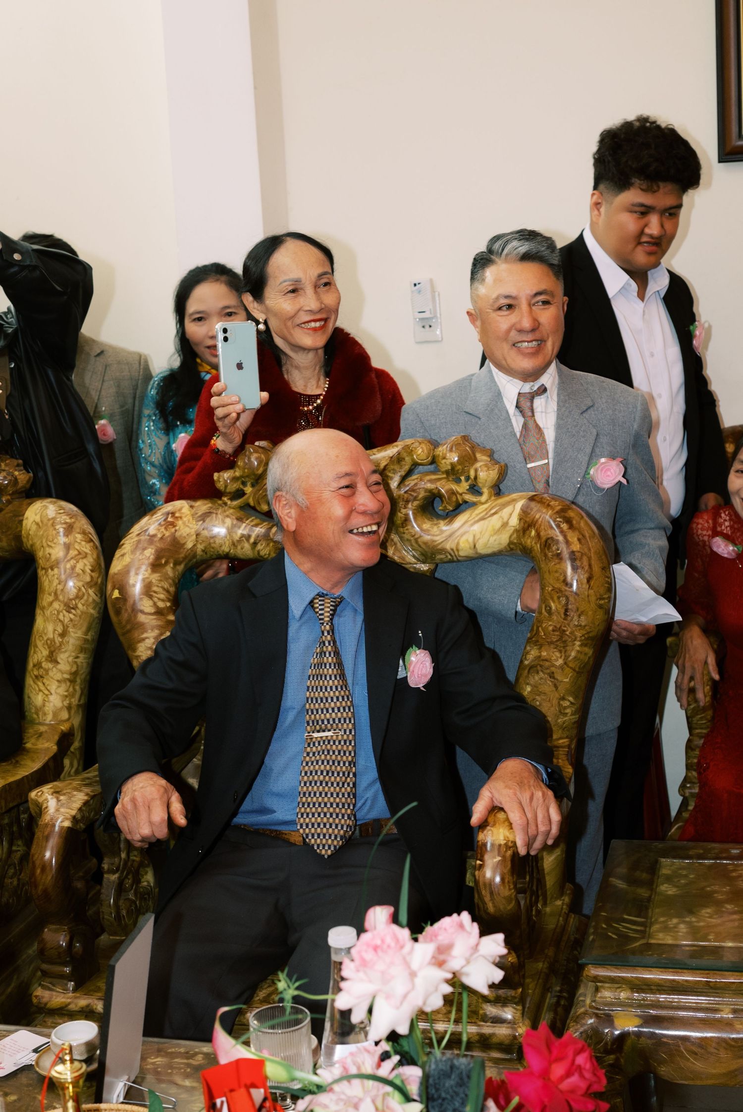 Elderly man in suit sits in ornate golden chair surrounded by smiling family at Asian wedding.