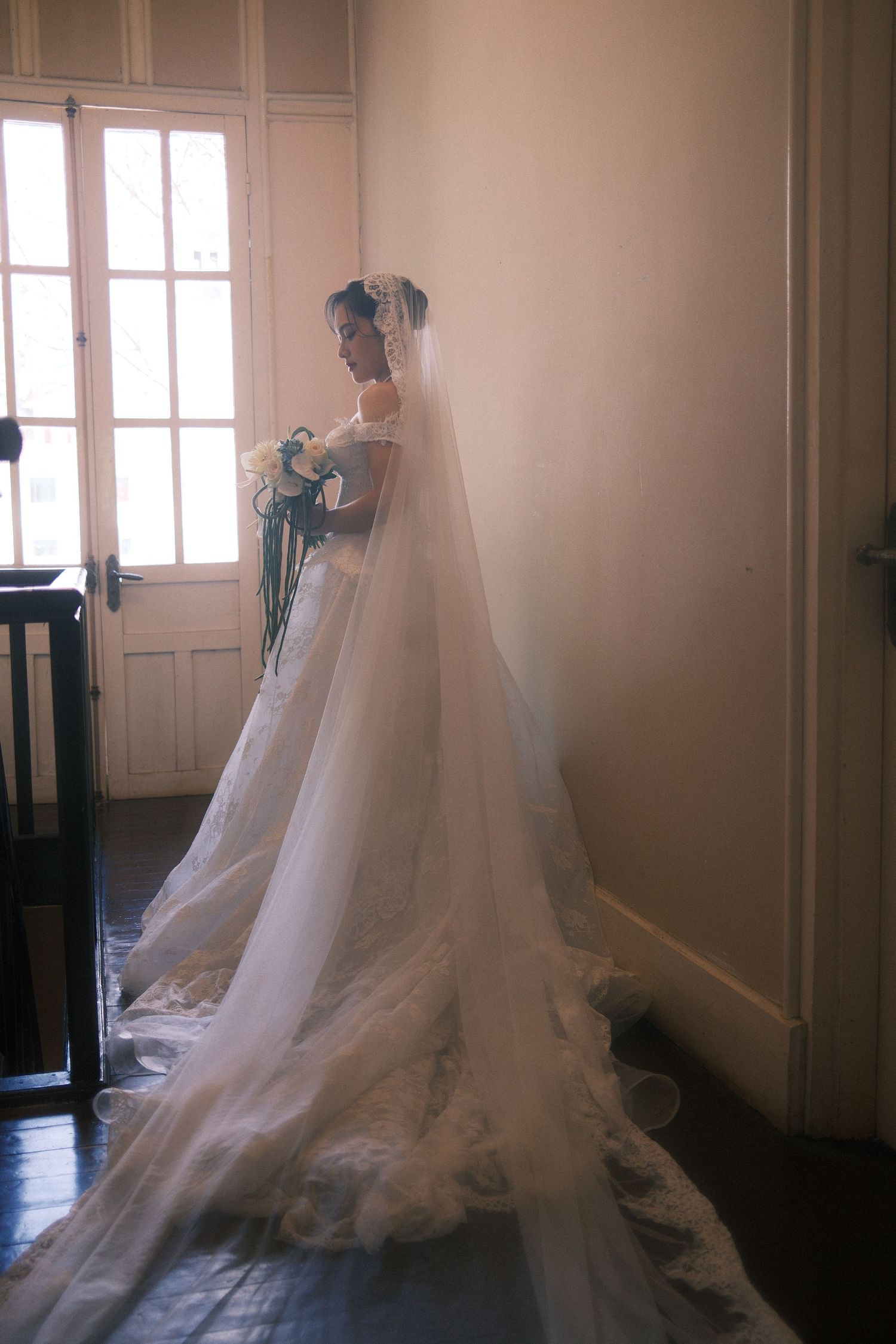 Bride in elegant white lace wedding gown with long veil holding bouquet, standing by window in soft natural light.