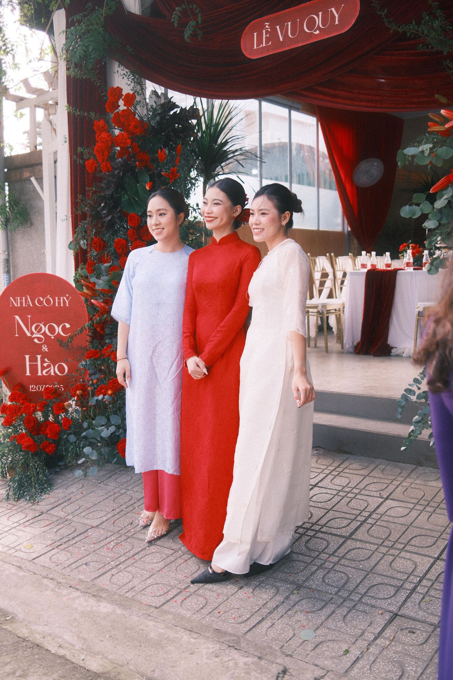 Three women in elegant ao dai dresses pose at a Vietnamese wedding ceremony decorated with red roses.