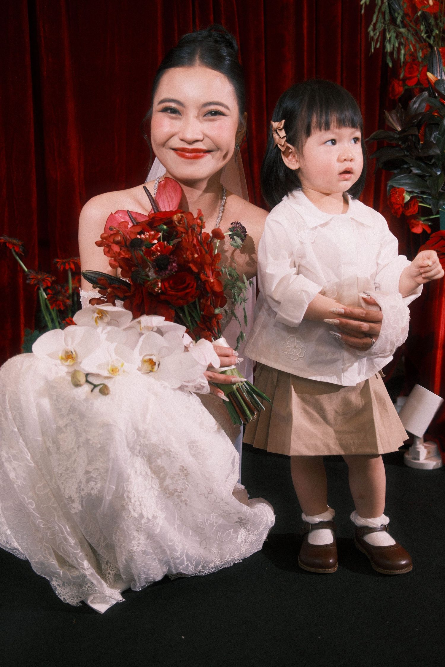 Bride in white lace gown holds red bouquet beside young toddler girl in beige outfit at wedding.
