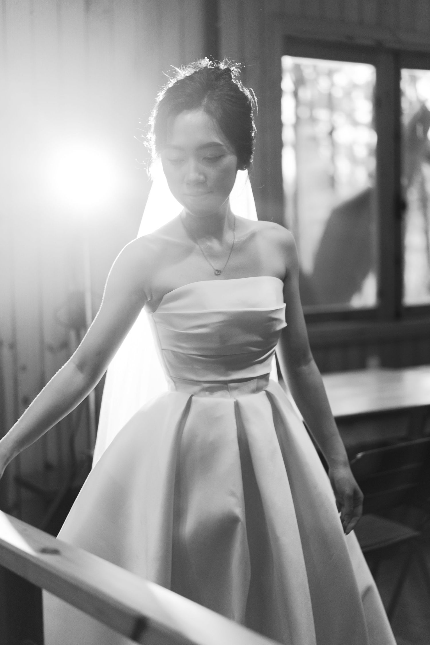 Black and white portrait of a bride in a strapless ball gown standing by a staircase railing with soft backlight.
