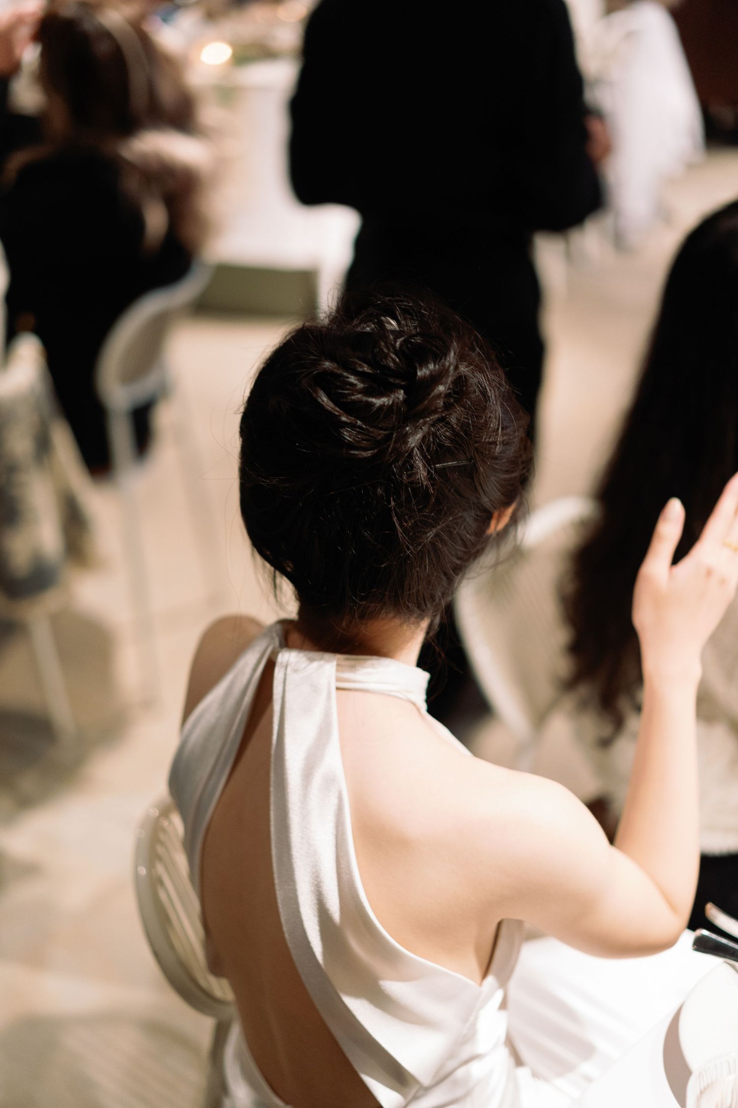 Back view of Asian woman in white halter dress with braided updo at indoor wedding reception.