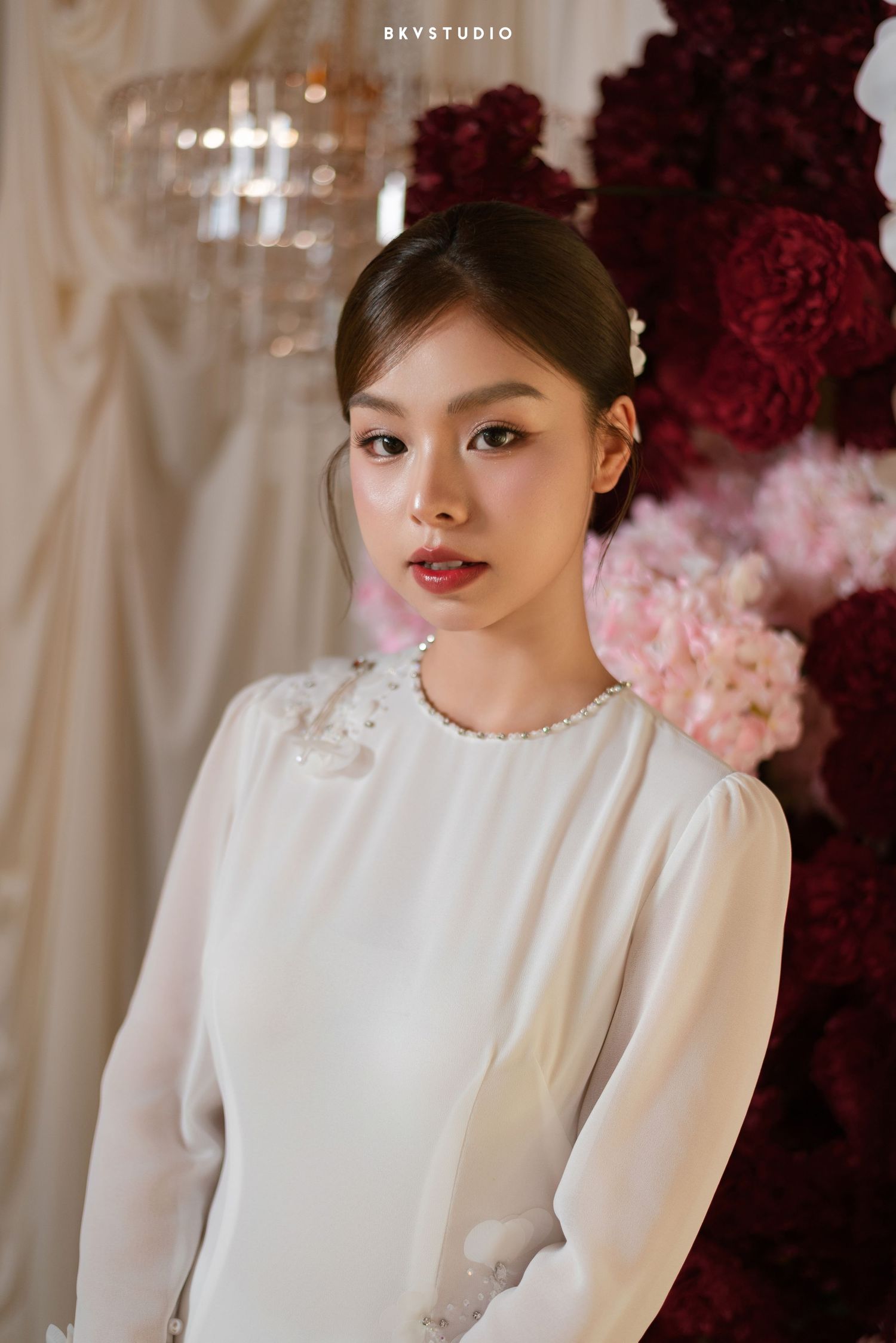 Young woman in white long-sleeve dress with pearl necklace poses before red and pink floral backdrop.