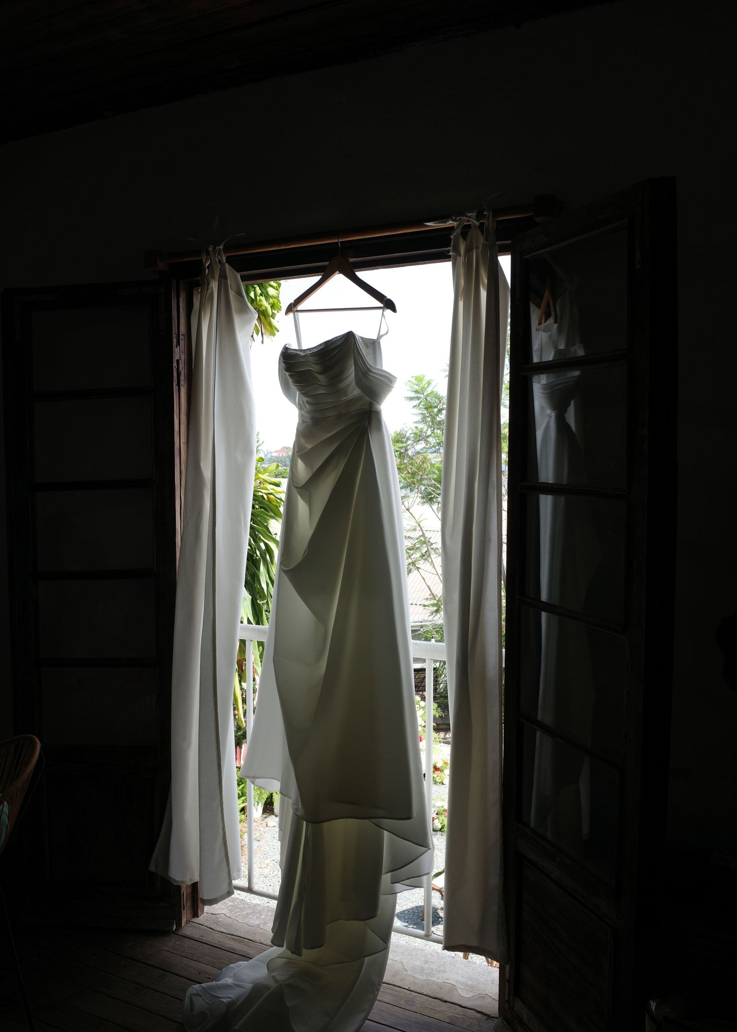 A white strapless wedding gown hanging on a hanger in a sunlit doorway with white curtains and lush greenery outside.