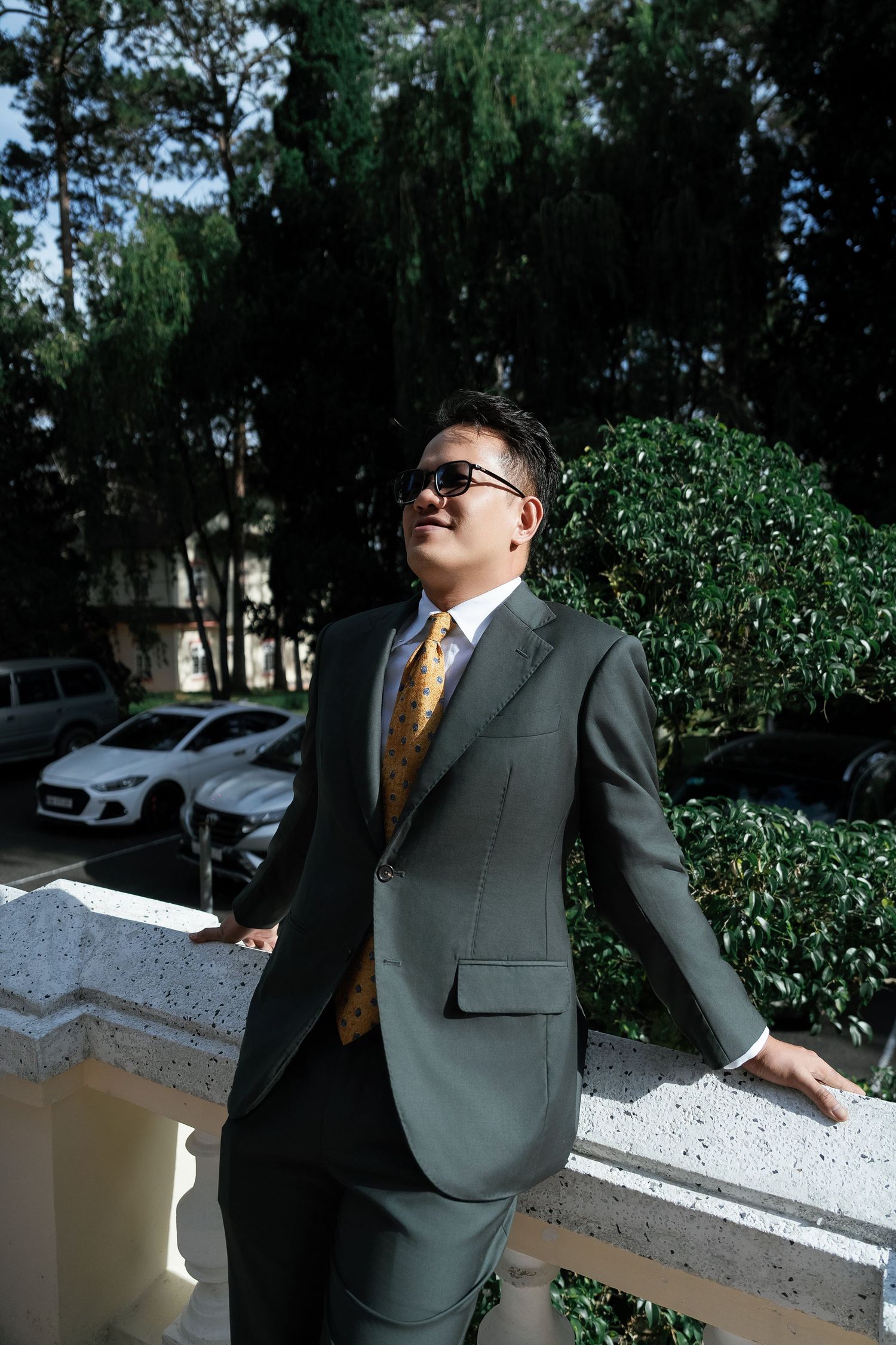 A man in a gray suit and gold tie wearing sunglasses stands on a balcony surrounded by lush greenery.