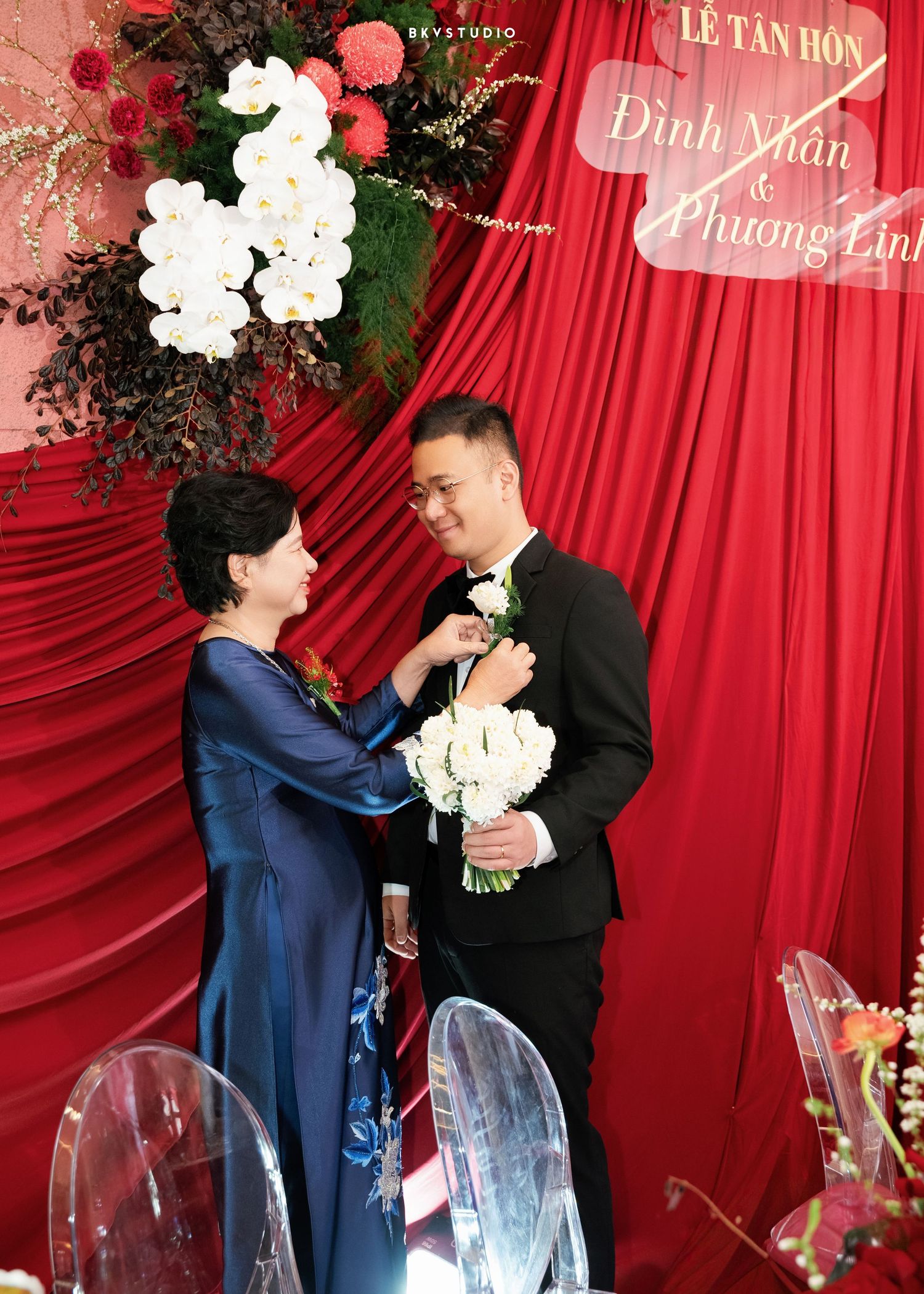 A groom in a black tuxedo receives a boutonniere from a woman in blue ao dai at a Vietnamese wedding ceremony.
