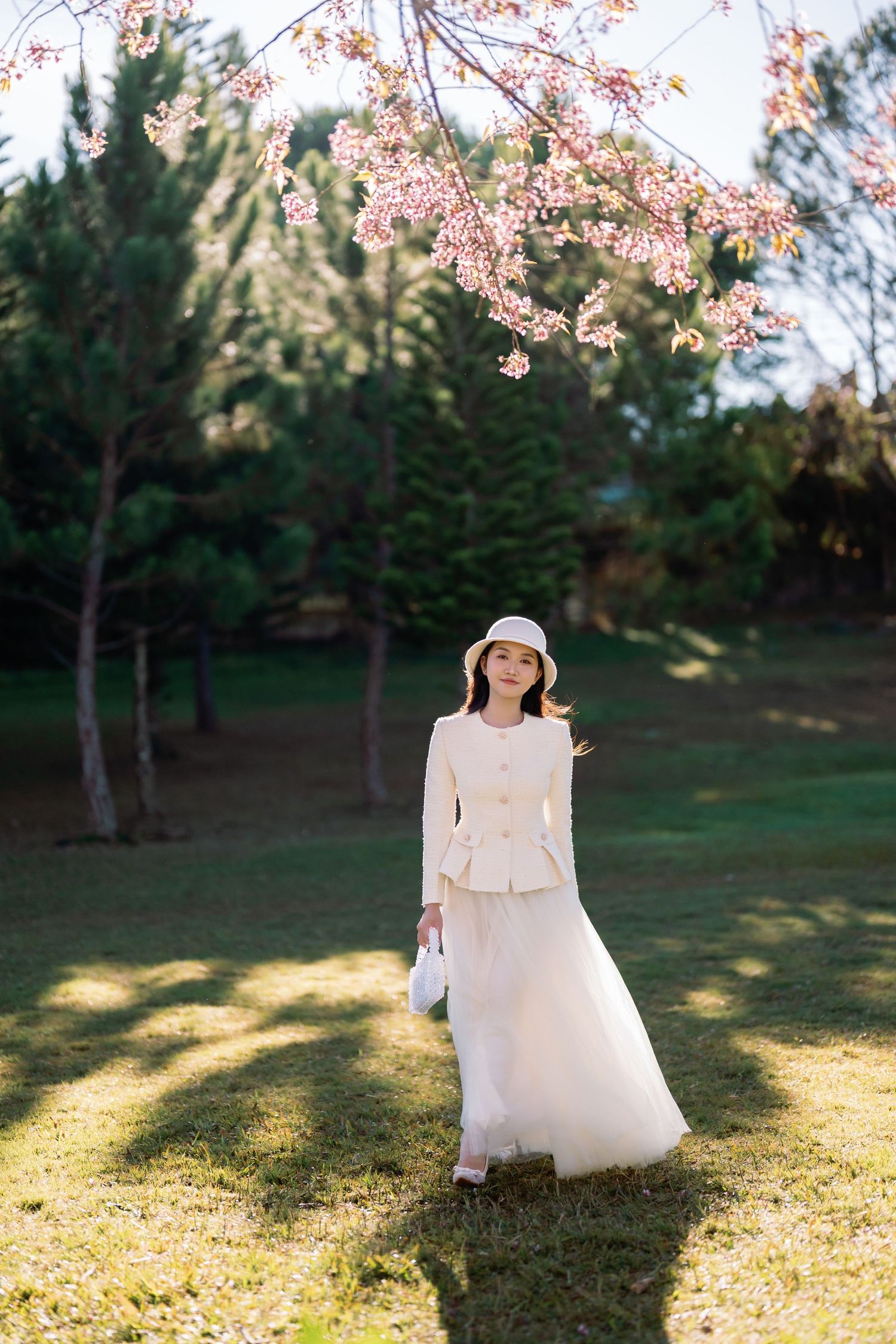 Woman in white jacket, skirt, and hat standing under pink cherry blossom tree in a sunny green park.