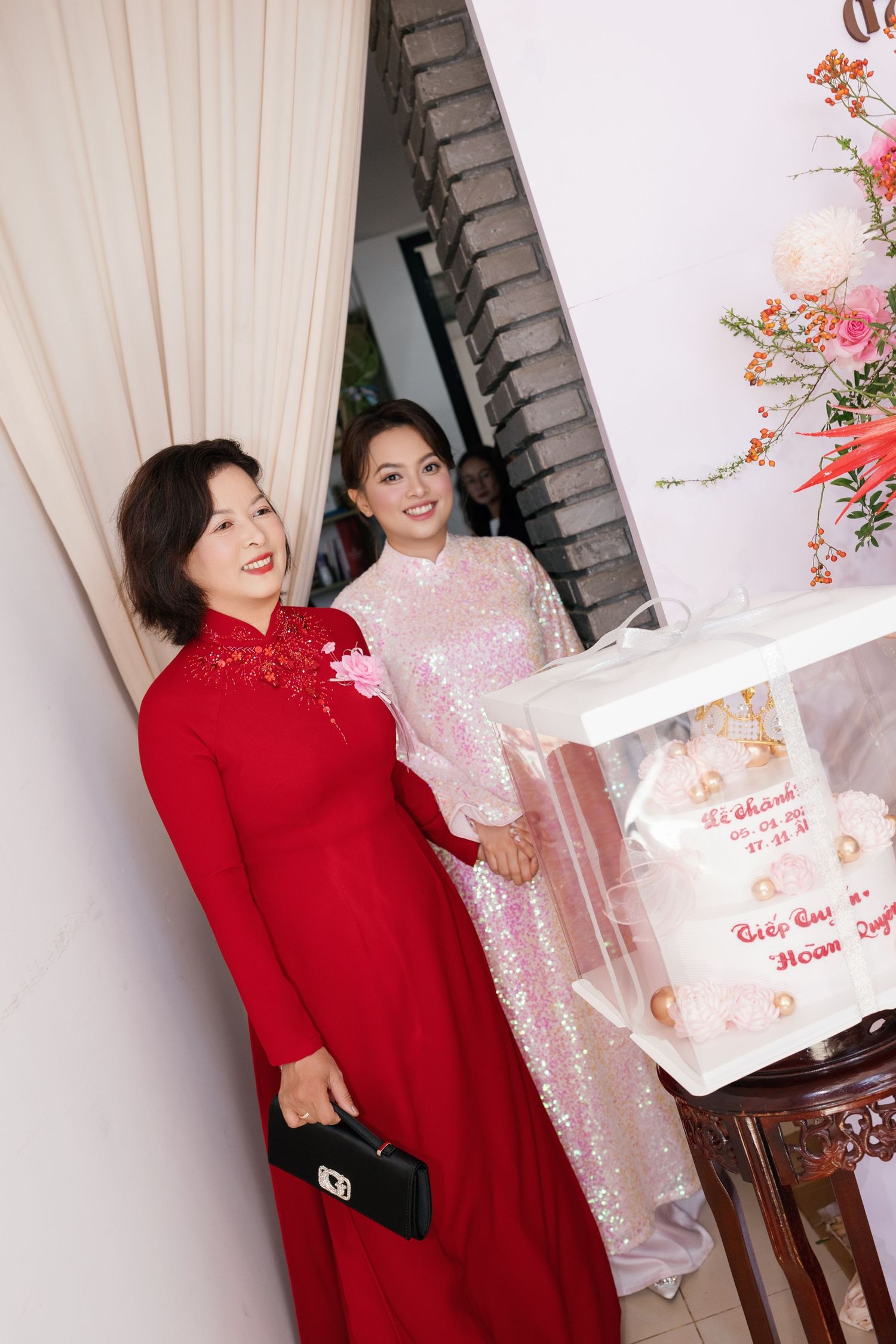 Two women in ao dai, red and pink sequined, pose together at Vietnamese wedding celebration.