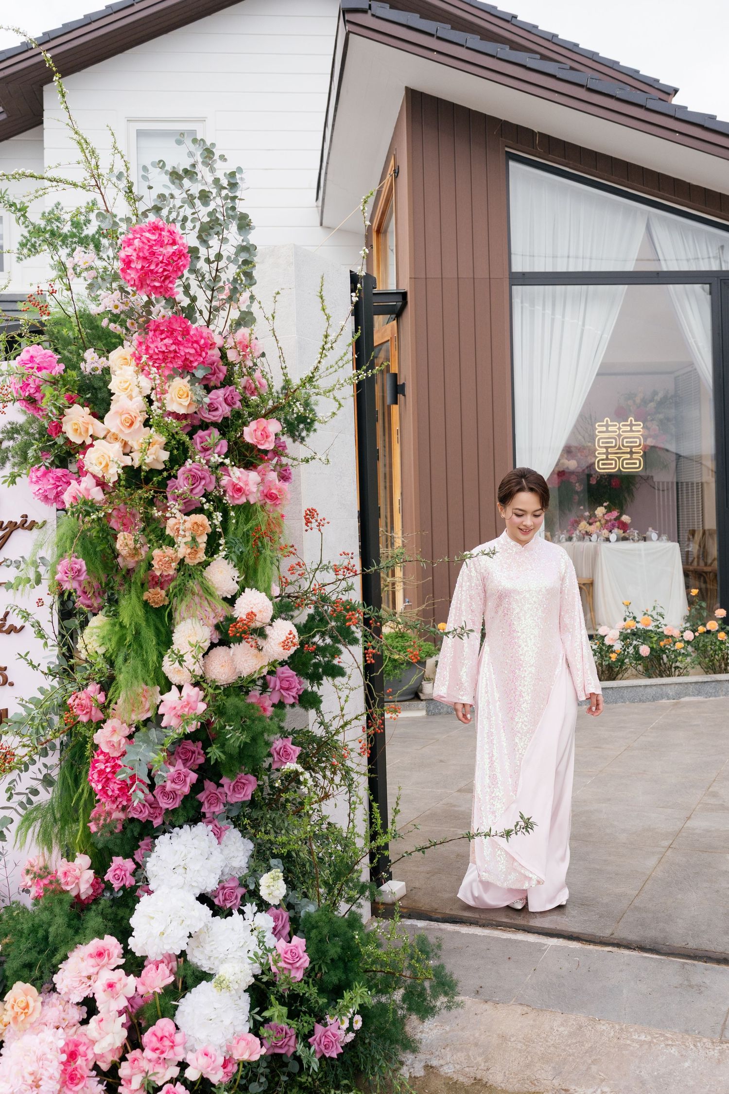 A bride in a pink floral ao dai walks past an elaborate pink and white floral wedding arch.
