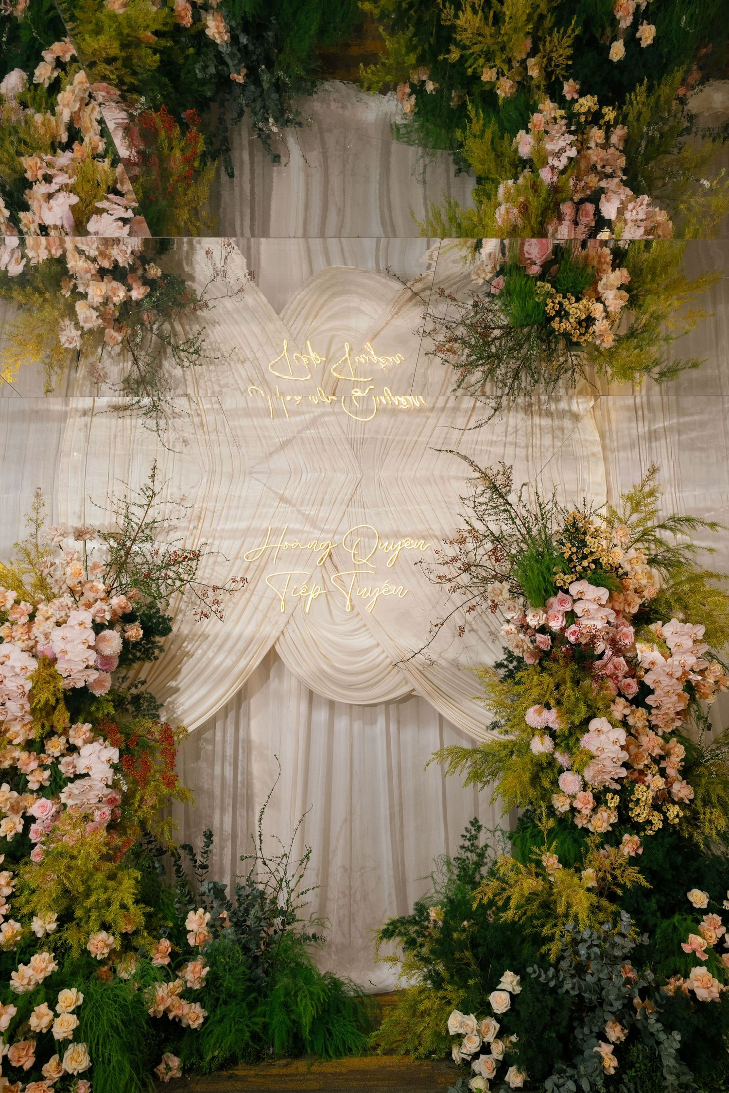 Romantic wedding backdrop with white draping, gold script signage, and lush floral arrangements of pink and yellow blooms.