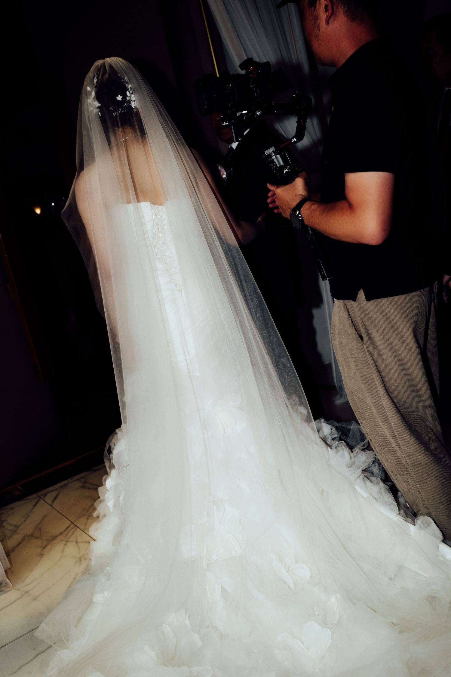 Bride in elegant white lace wedding gown with long veil being photographed by cameraman.