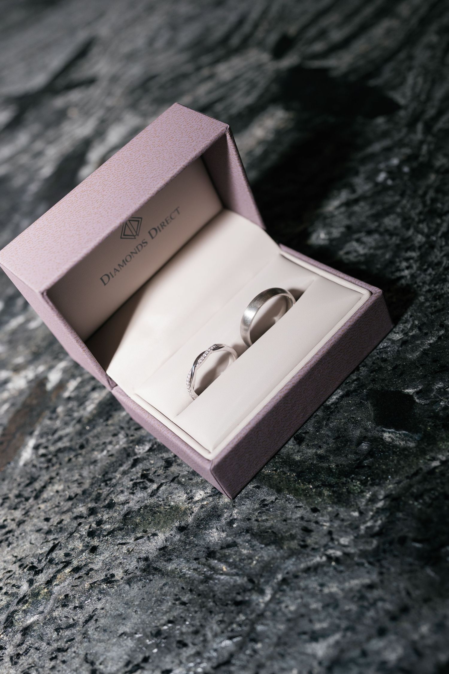 Two silver wedding bands displayed in an open pink Diamond Direct jewelry box on a dark granite surface.