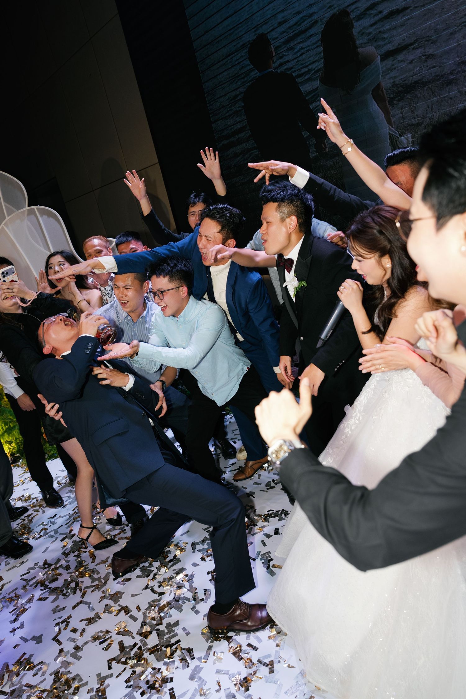 Groom and groomsmen dancing energetically with bride at lively wedding reception celebration with confetti on floor.