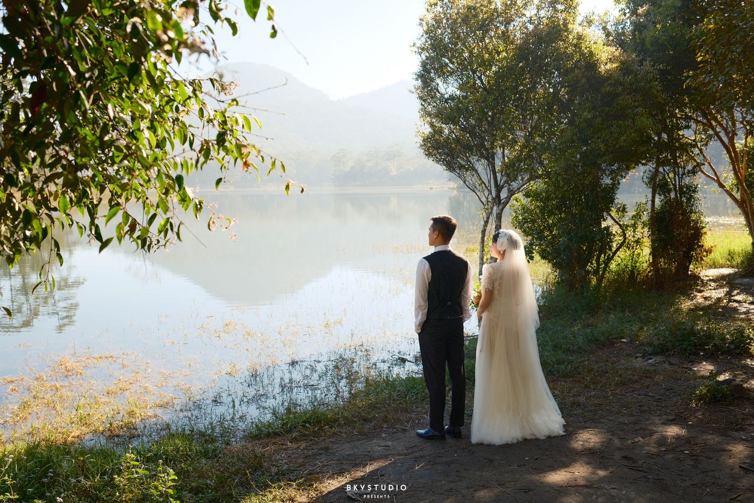 Bride and groom stand together facing a serene reflective lake surrounded by trees and misty hills.