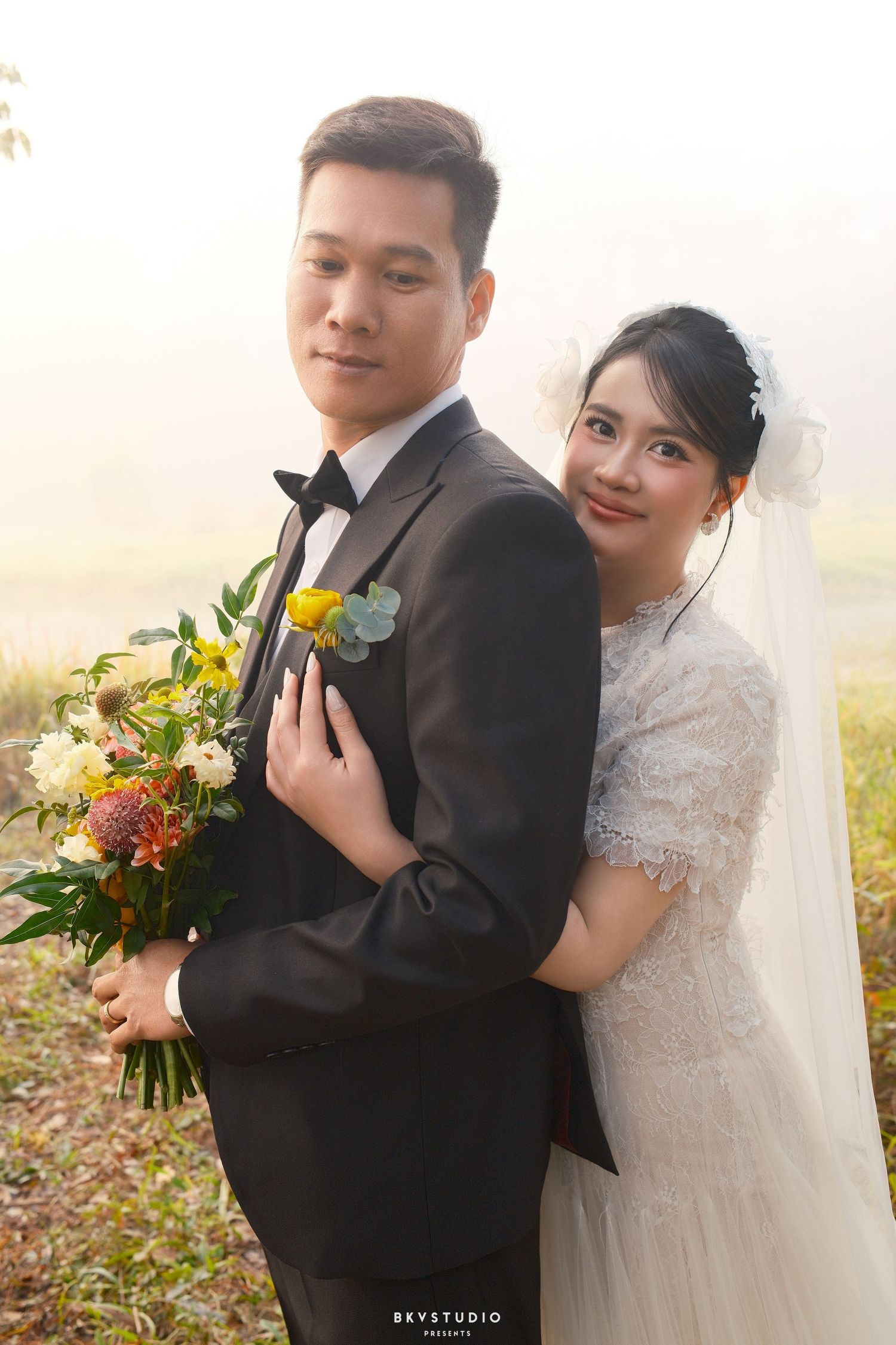 Bride in white lace wedding dress embraces groom in black suit from behind, holding a vibrant floral bouquet outdoors.
